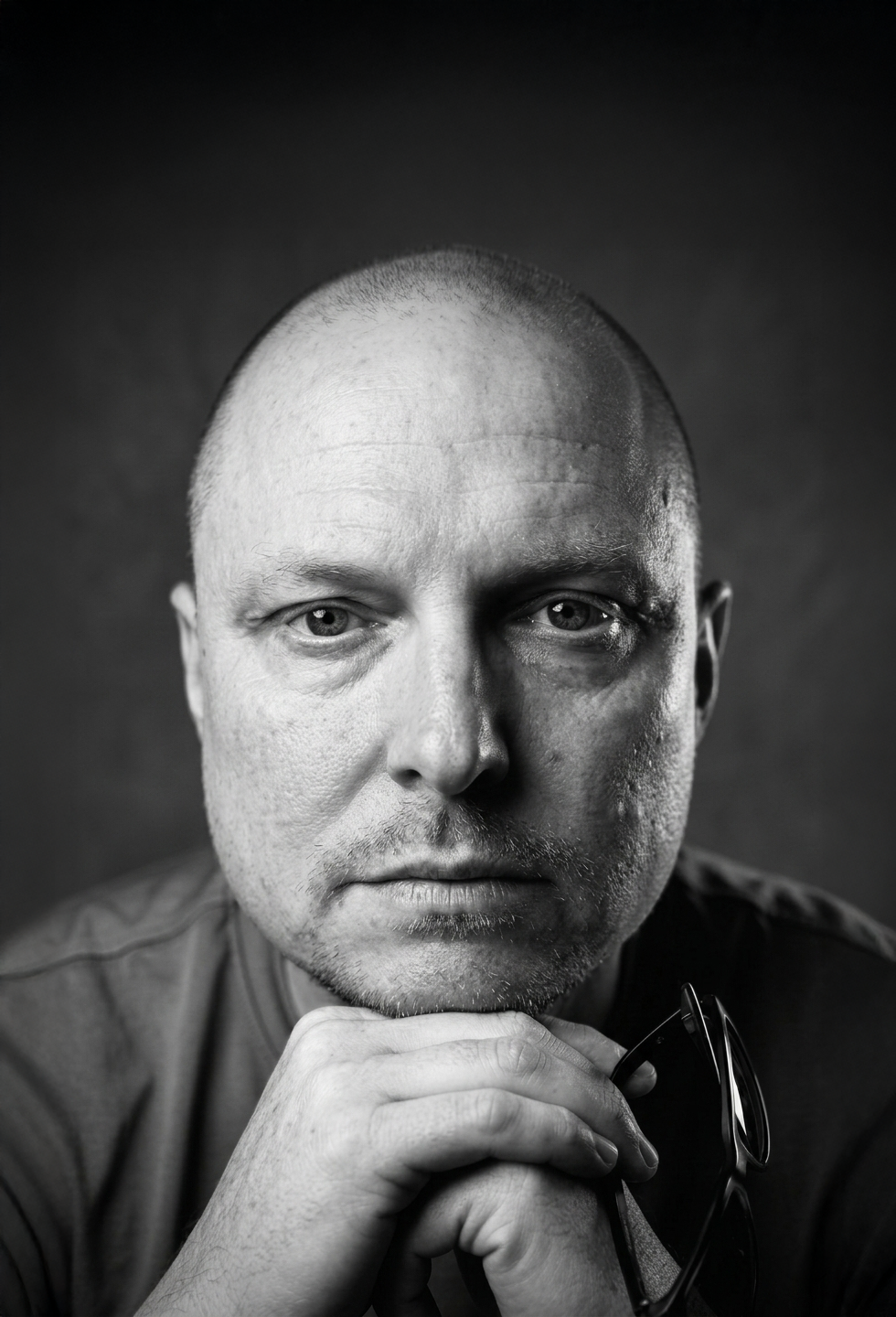 Black and white close-up portrait of a bald man resting his chin on his hand, holding sunglasses, against a dark background.