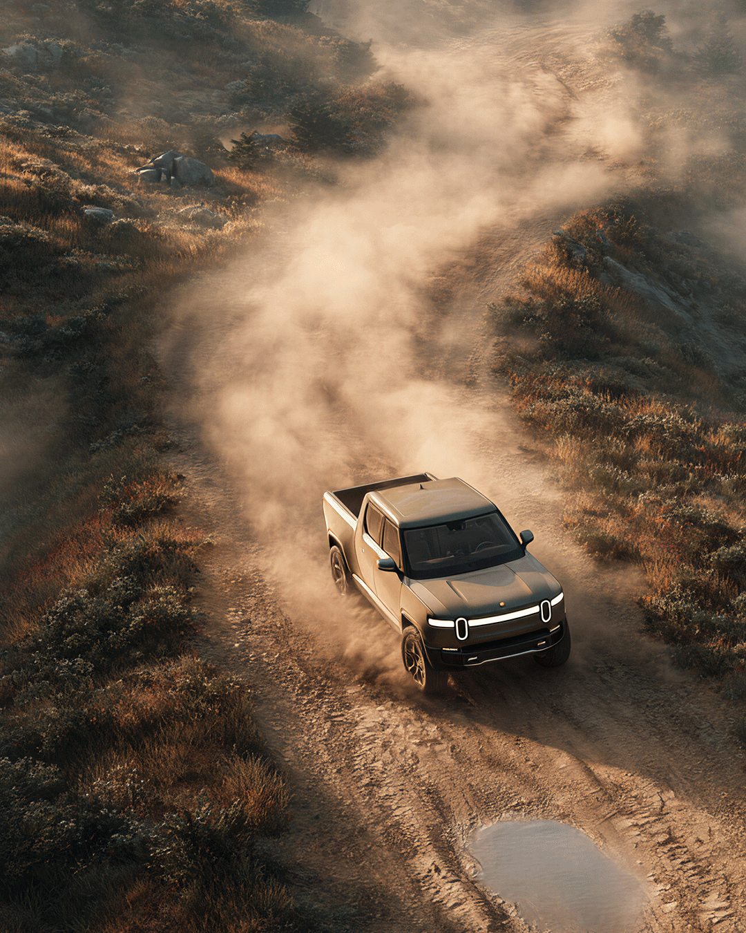 An electric pickup truck driving on a dirt road through a rugged landscape with dust clouds behind it and a puddle in front, with hills and vegetation in the background.