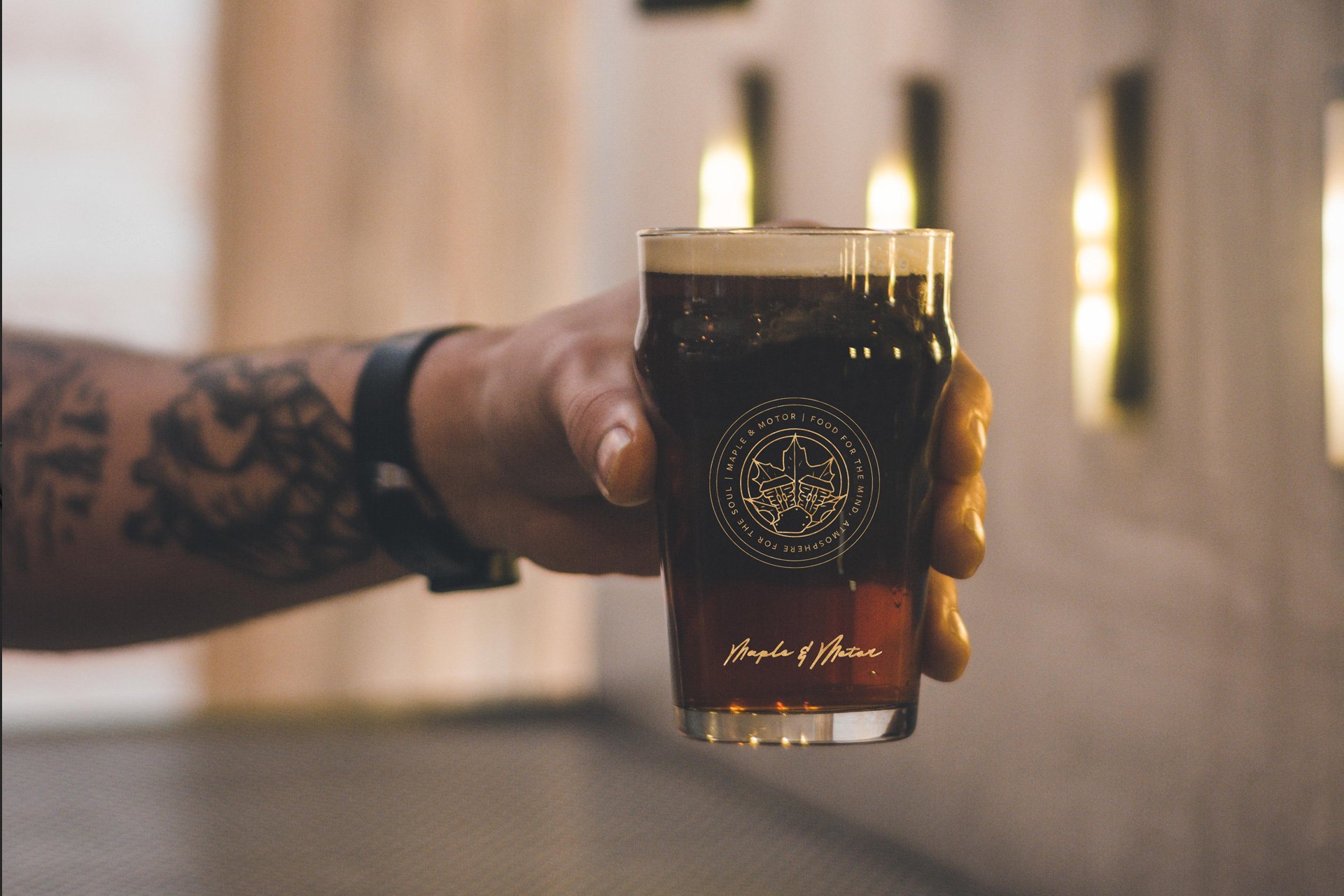 A person with a tattooed arm holding a glass of dark cola, with a blurred background featuring vertical lights.