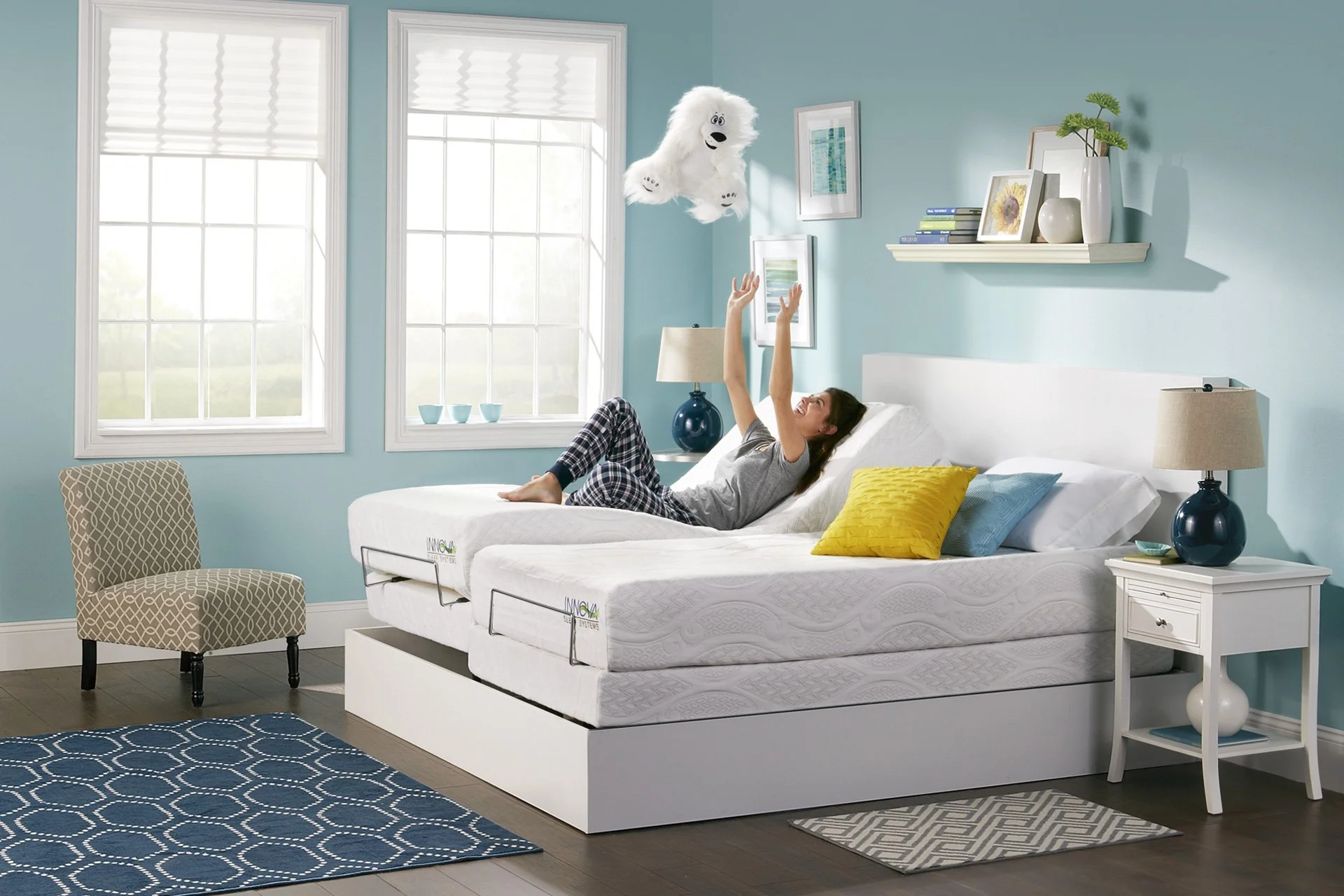 A woman lying on a bed in a light blue bedroom, tossing a white fluffy stuffed animal into the air. There are two large windows behind her, a chair on the left, and two nightstands with lamps on each side of the bed. The room has a blue and white col