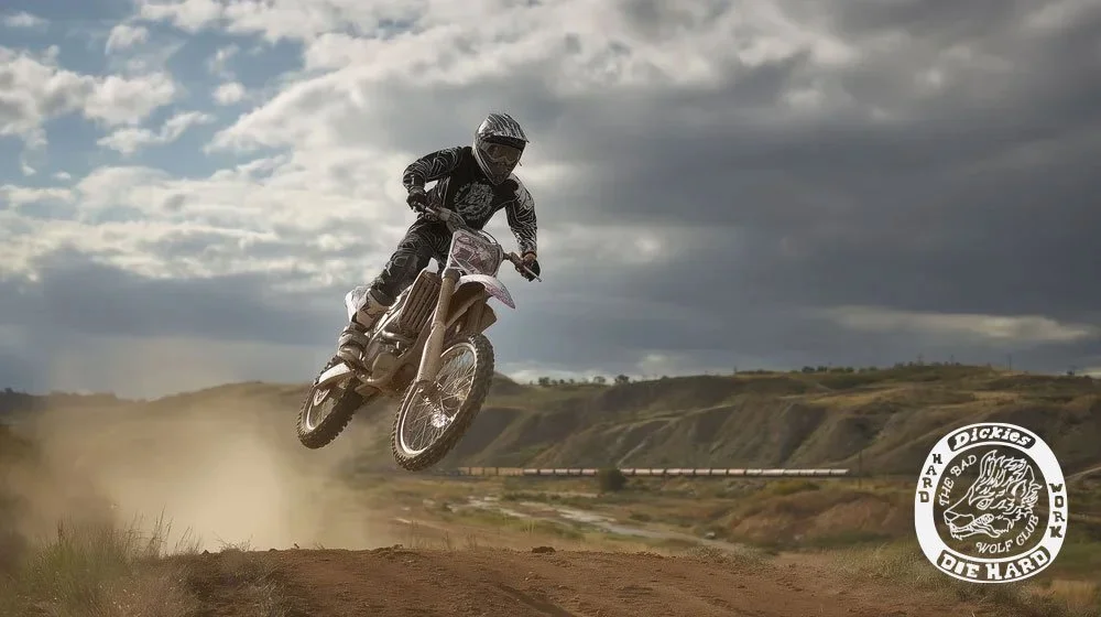 A person in motocross gear jumps a dirt bike off a ramp on a dirt track with a cloudy sky overhead.