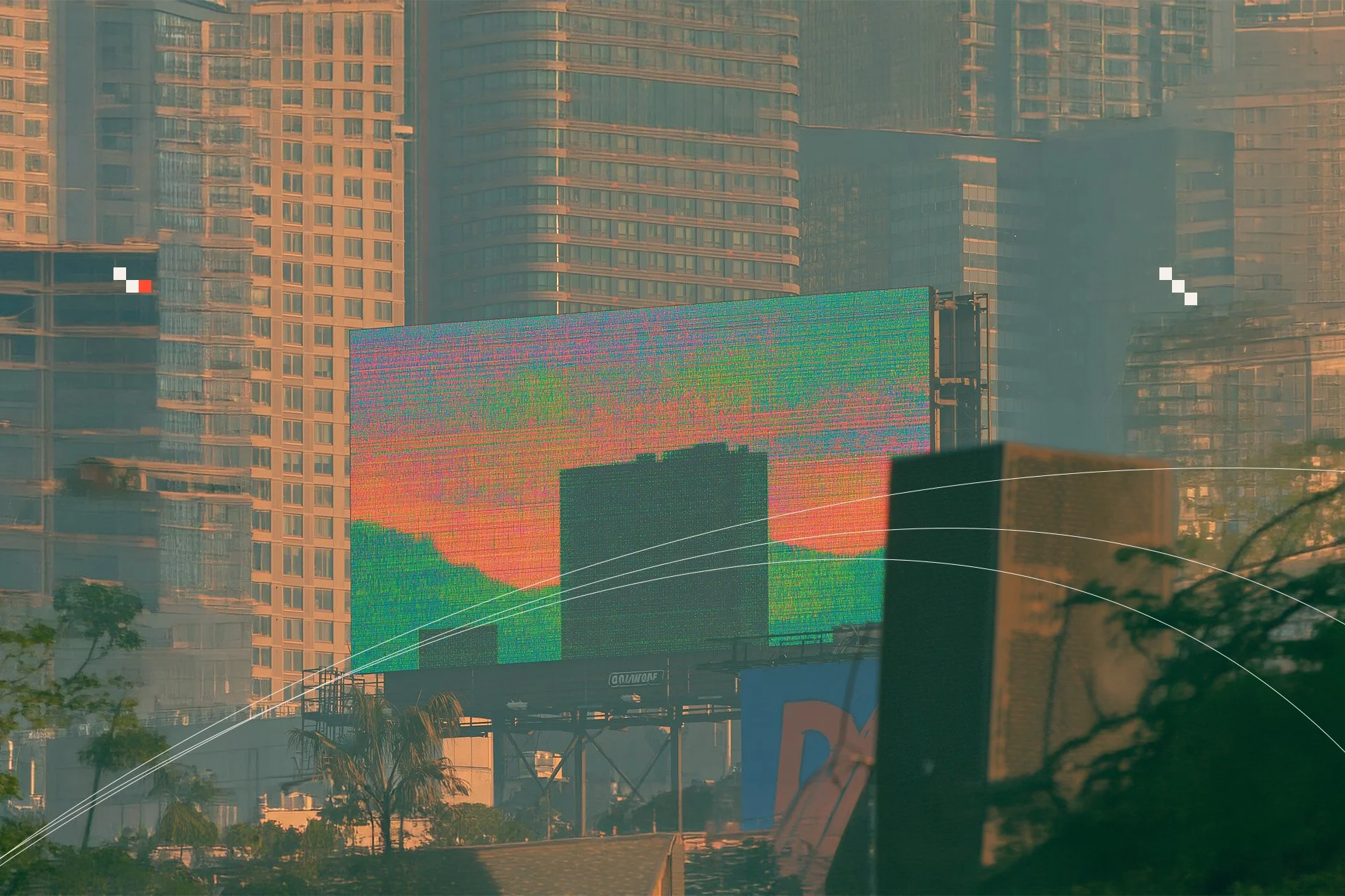 Cityscape featuring tall modern glass buildings with a large digital billboard displaying colorful static, set against an orange-tinted sky during sunset or sunrise, with trees and a vehicle window reflection in the foreground.