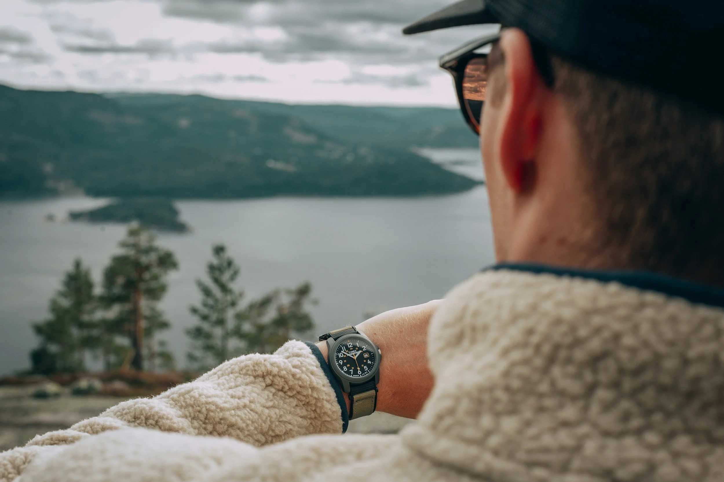 A person wearing sunglasses and a fleece jacket looking out over a lake with hills in the background, checking a watch.