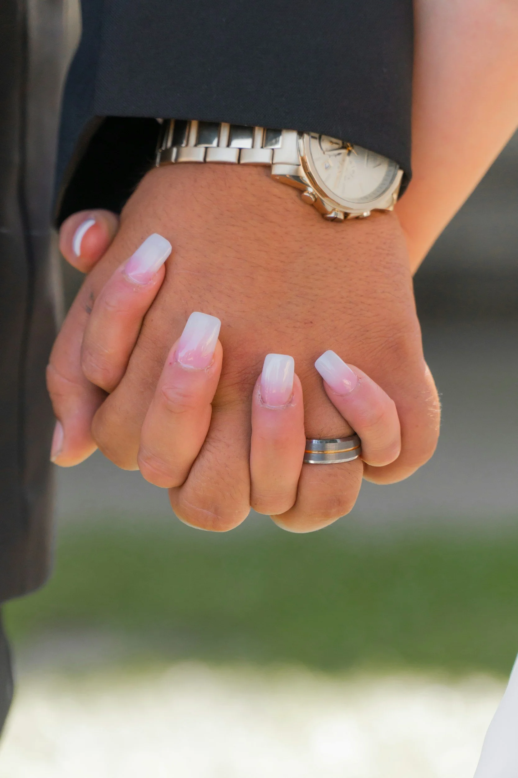 Close-up of a man's hand wearing a silver watch and wedding band, interlocked with a woman's hand with long, manicured nails, also wearing a wedding band.