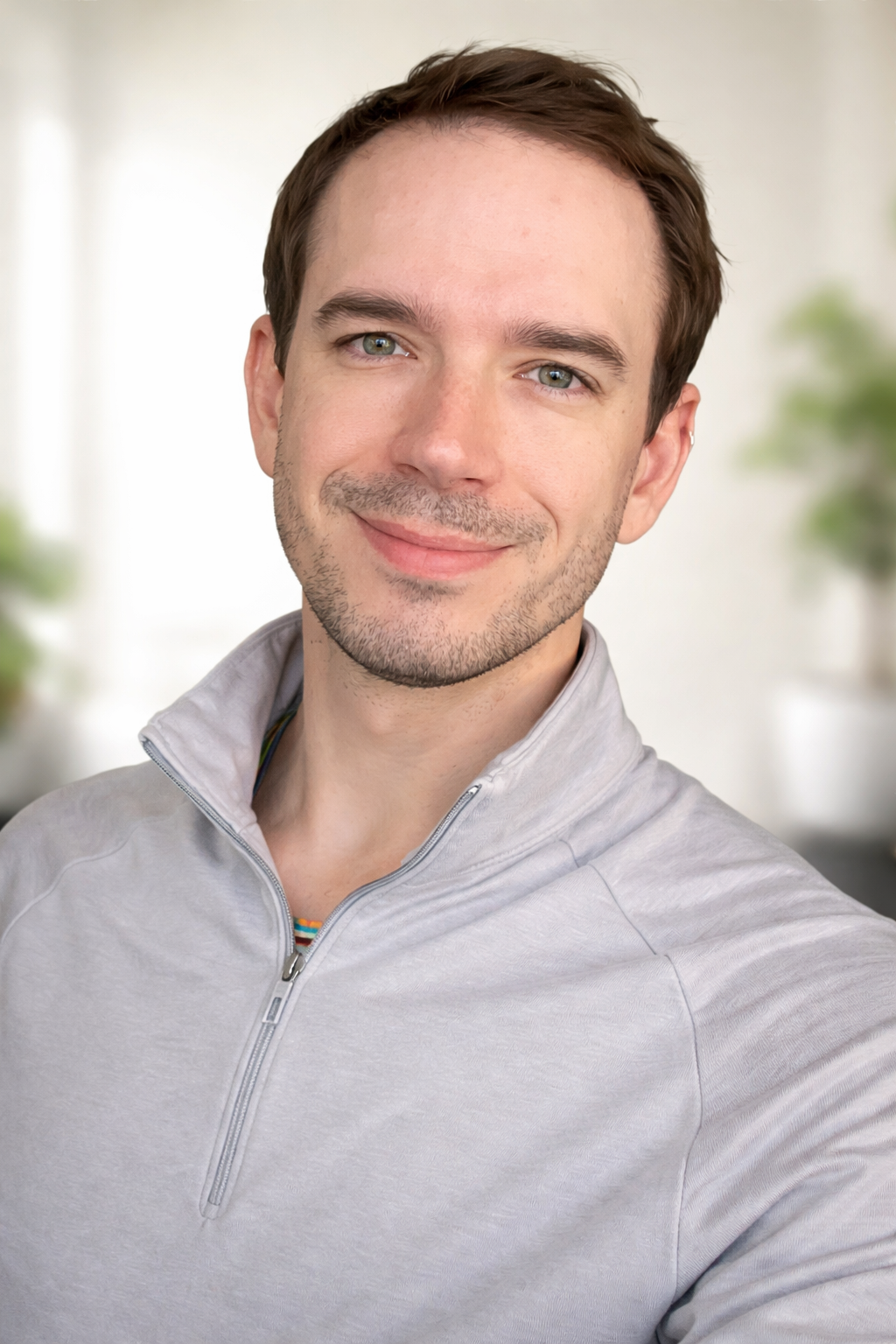 A smiling young man with short brown hair and blue eyes, wearing a light gray zip-up jacket, standing indoors with a blurred background of plants and bright light.