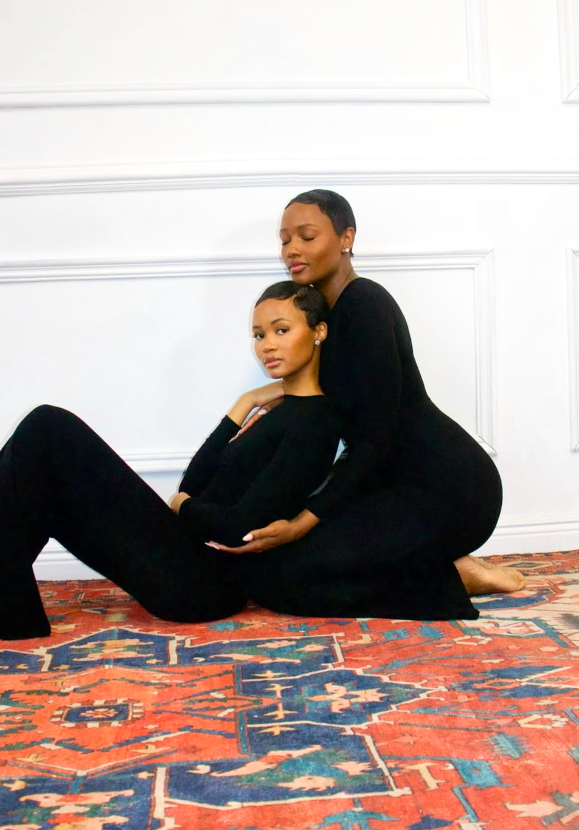 Two women in black outfits pose on a patterned rug against a white paneled wall. One woman is seated on the floor with her arm around the other woman, who is kneeling behind her, both with calm expressions and short hairstyles.