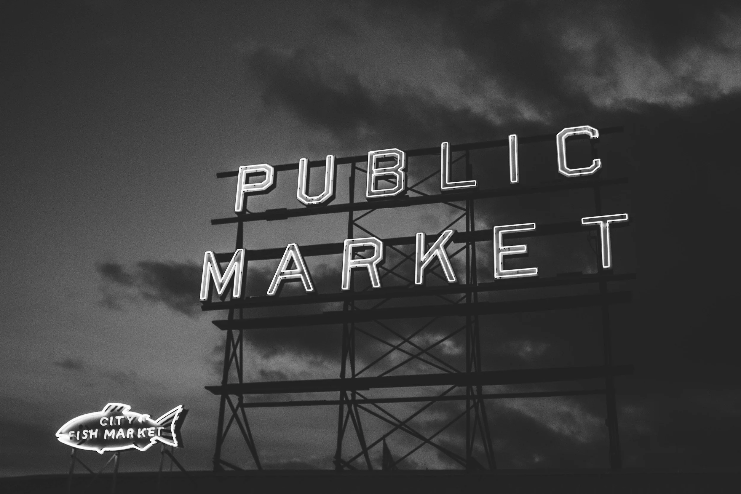 Neon sign reading 'Public Market' with a smaller sign below in the shape of a fish that reads 'City Fish Market', set against a cloudy sky at dusk.