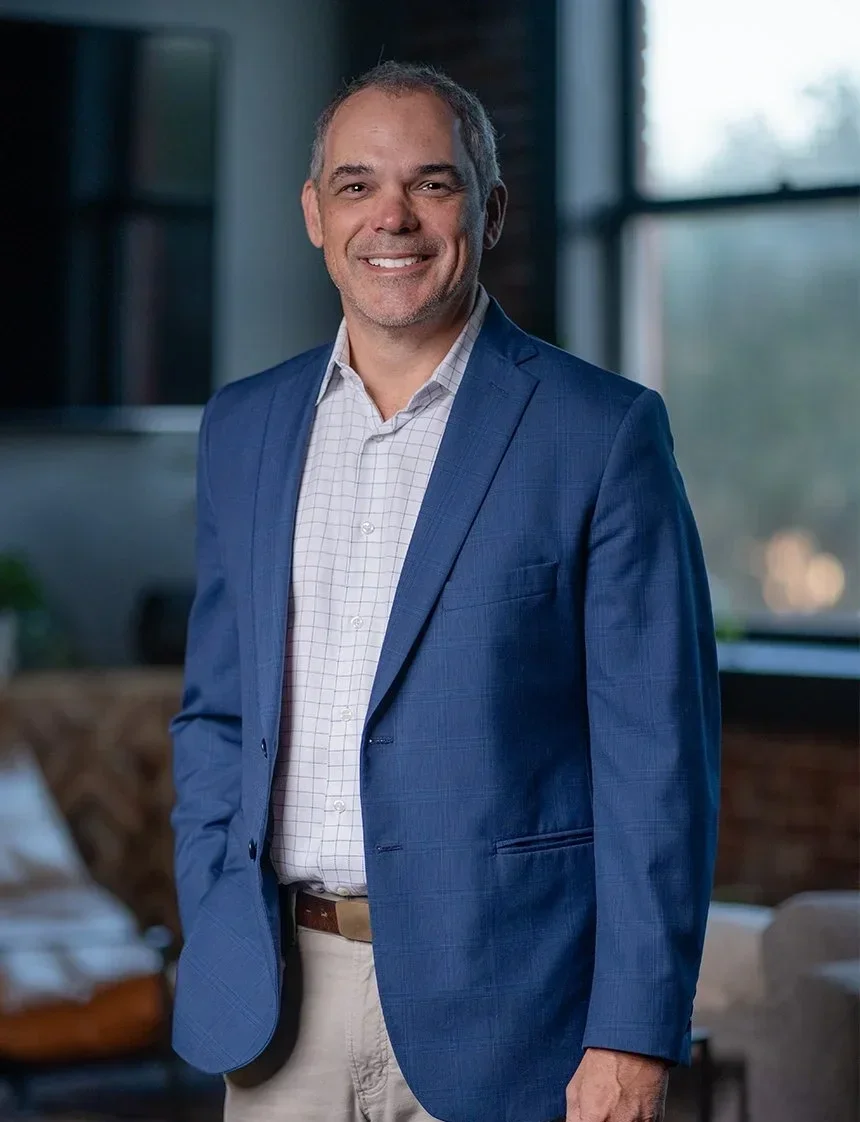 A middle-aged man with short hair, wearing a blue blazer over a white checkered shirt, smiling, standing indoors near large windows.