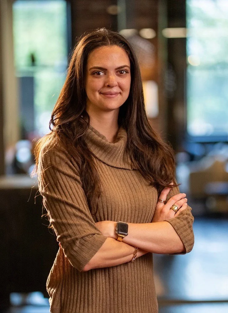 A woman with long dark hair, wearing a brown ribbed turtleneck sweater, standing indoors with arms crossed, smiling at the camera.