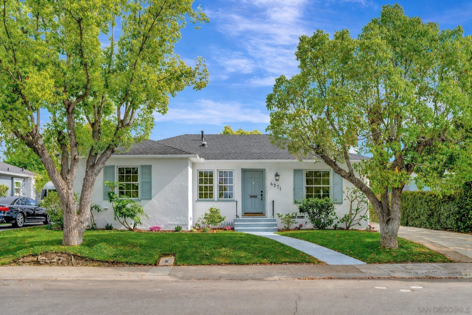 White house with light blue shutters and front door, two large trees flanking the house, well-kept lawn, flowers along the house, sidewalk in front, parked cars to the left, clear blue sky.