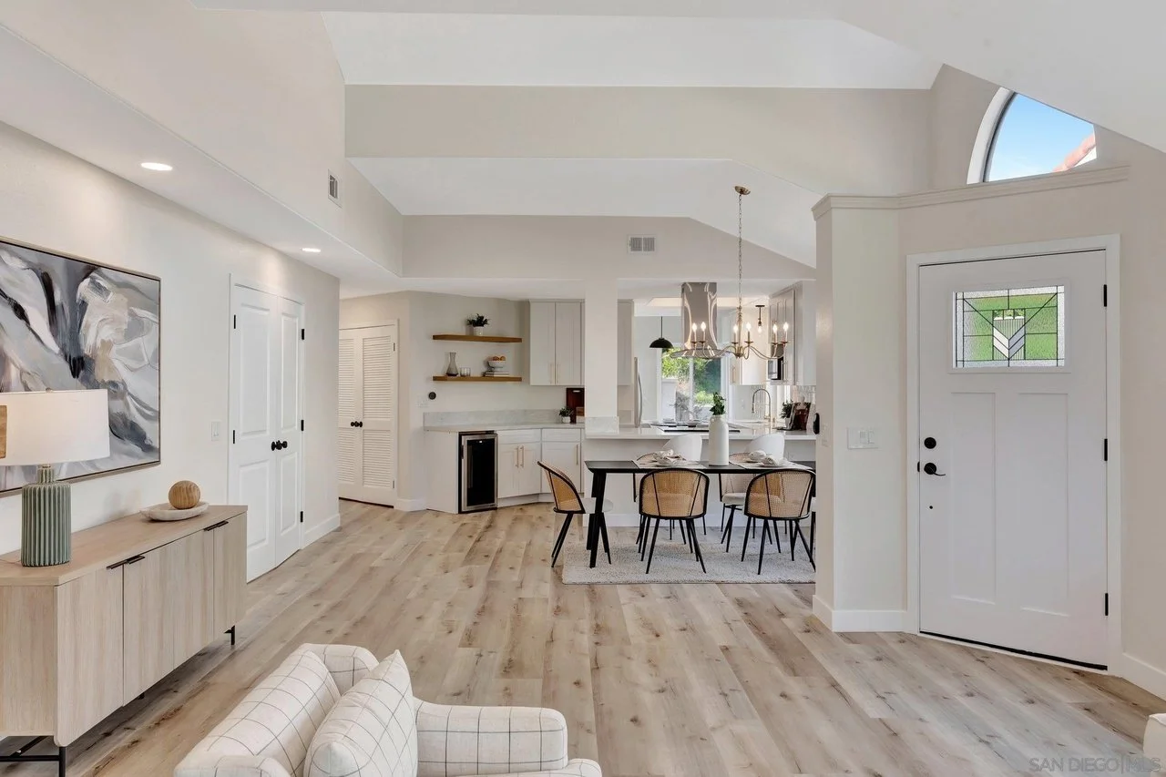 Modern open-concept living space with white walls, wood flooring, a dining area with a black table and six chairs, a kitchen with white cabinets, a chandelier above the dining table, and a front door with stained glass window.