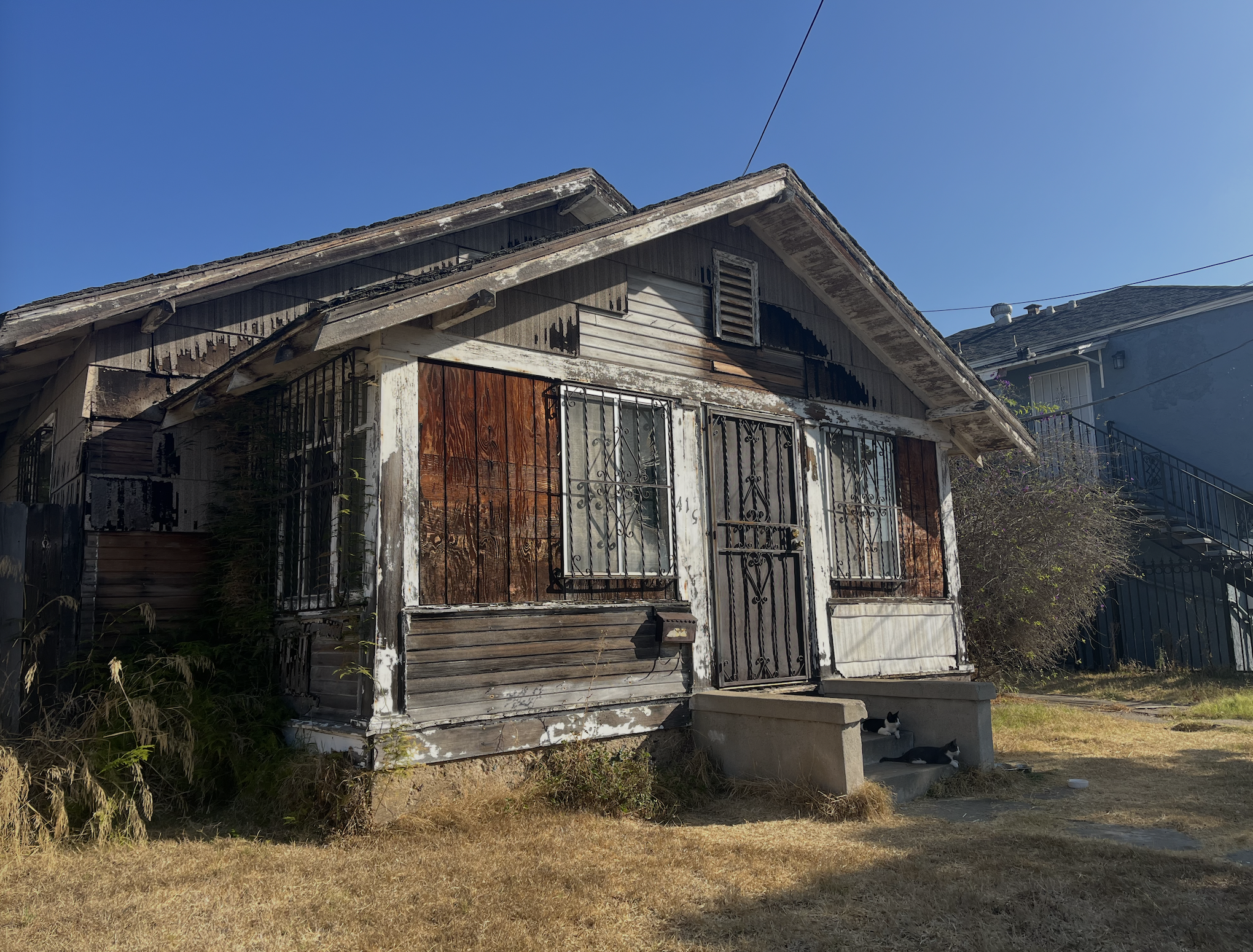 Room under renovation with exposed cabinetry, construction debris, and tools on the floor. Partially demolished wall and open doorway leading to a bright room with a door and windows. Ceiling fan and beams visible.