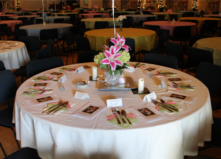 Round banquet table decorated with a floral centerpiece with pink lilies and white flowers, set with plates, gold cutlery, pink napkins, programs, candles, and name cards, in a large decorated event hall.