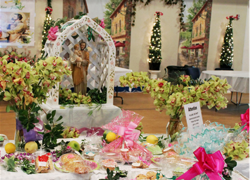 Display of religious figurines and floral arrangements with pink and purple ribbons, in a decorated indoor setting.