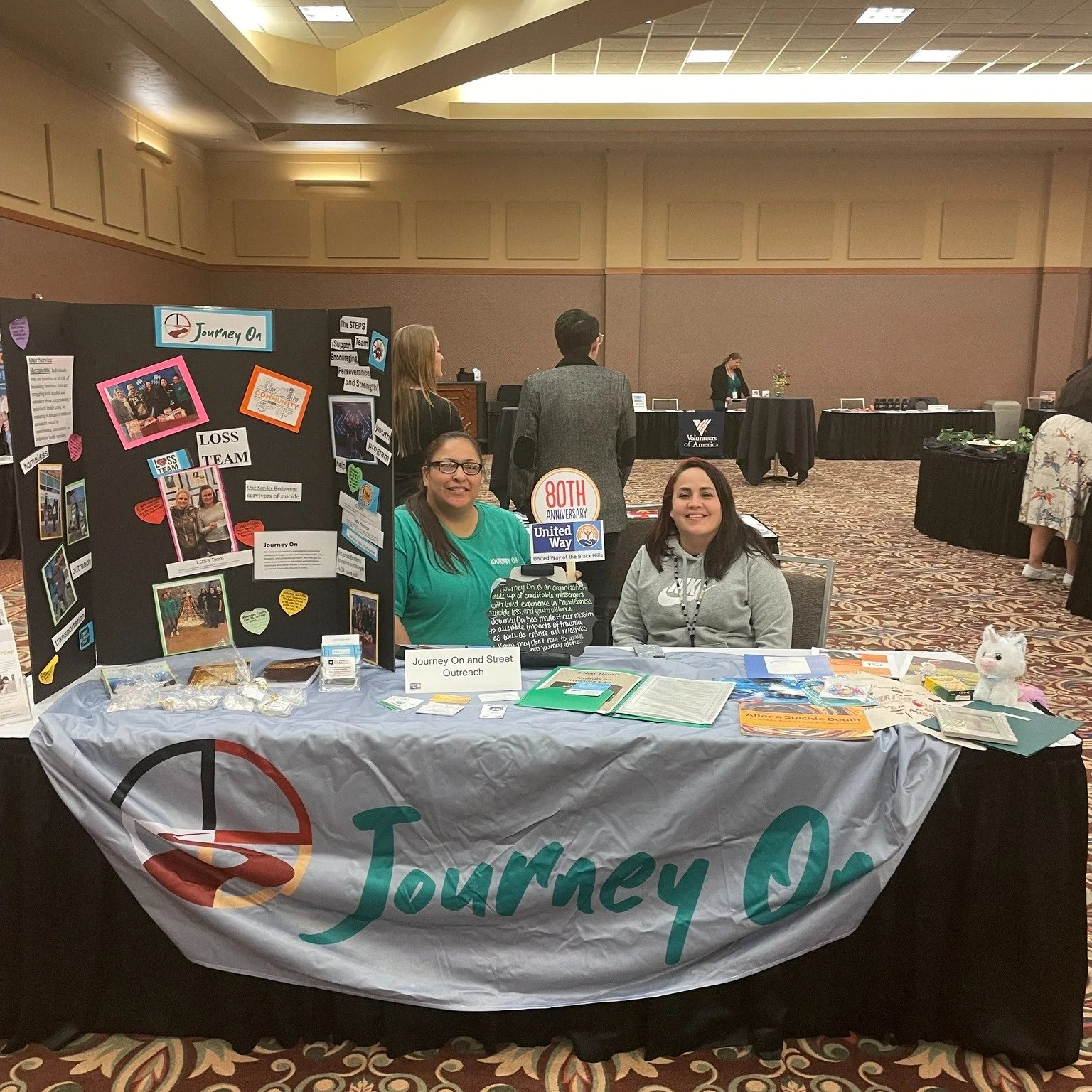 Two women sitting at a table with a Journey On banner, informational materials, and a display board about outreach and community support at a conference or event.