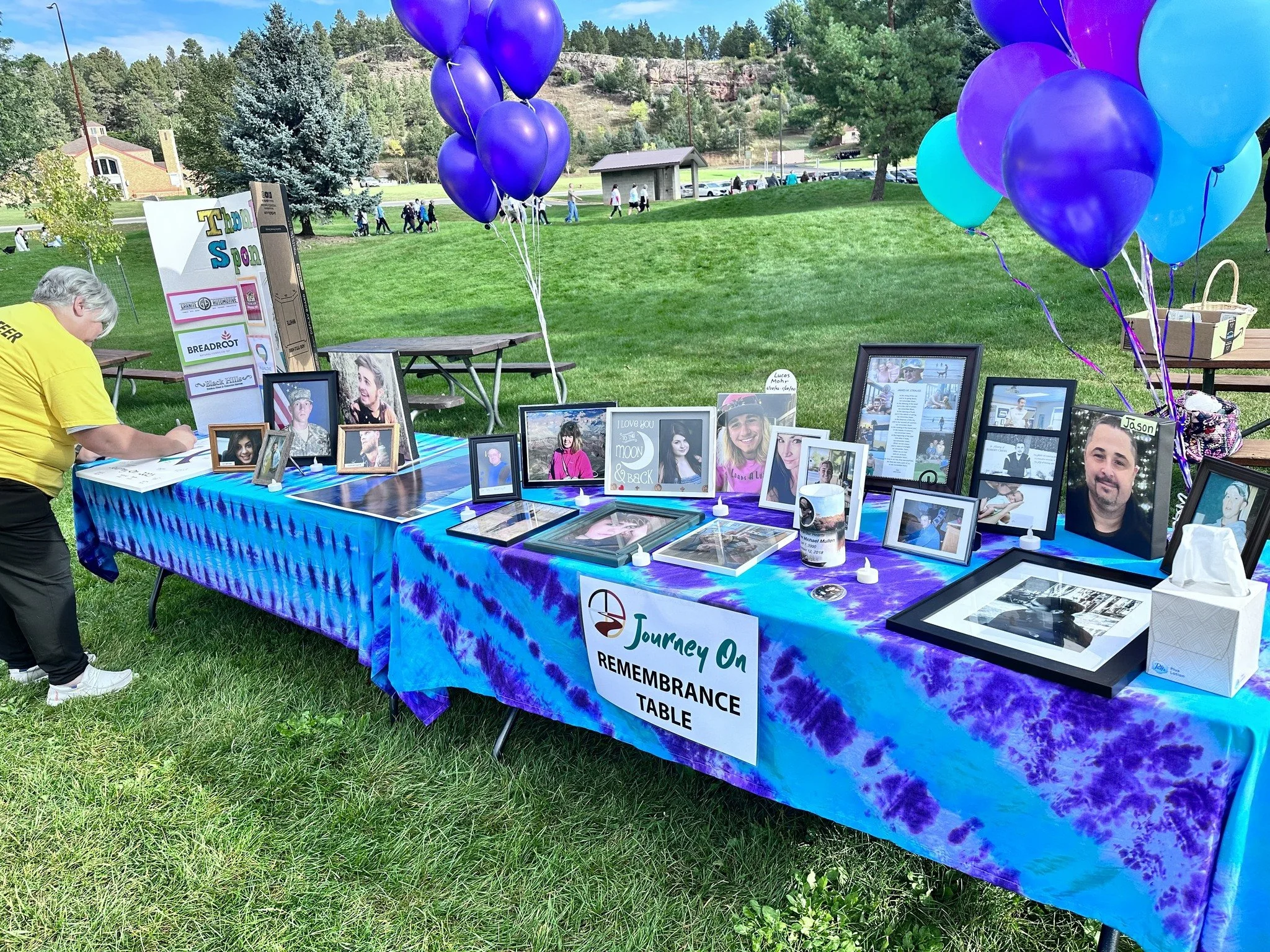A remembrance table at an outdoor event decorated with framed photographs of loved ones, purple and blue balloons, and a colorful tie-dye tablecloth, with a person signing a guest book.