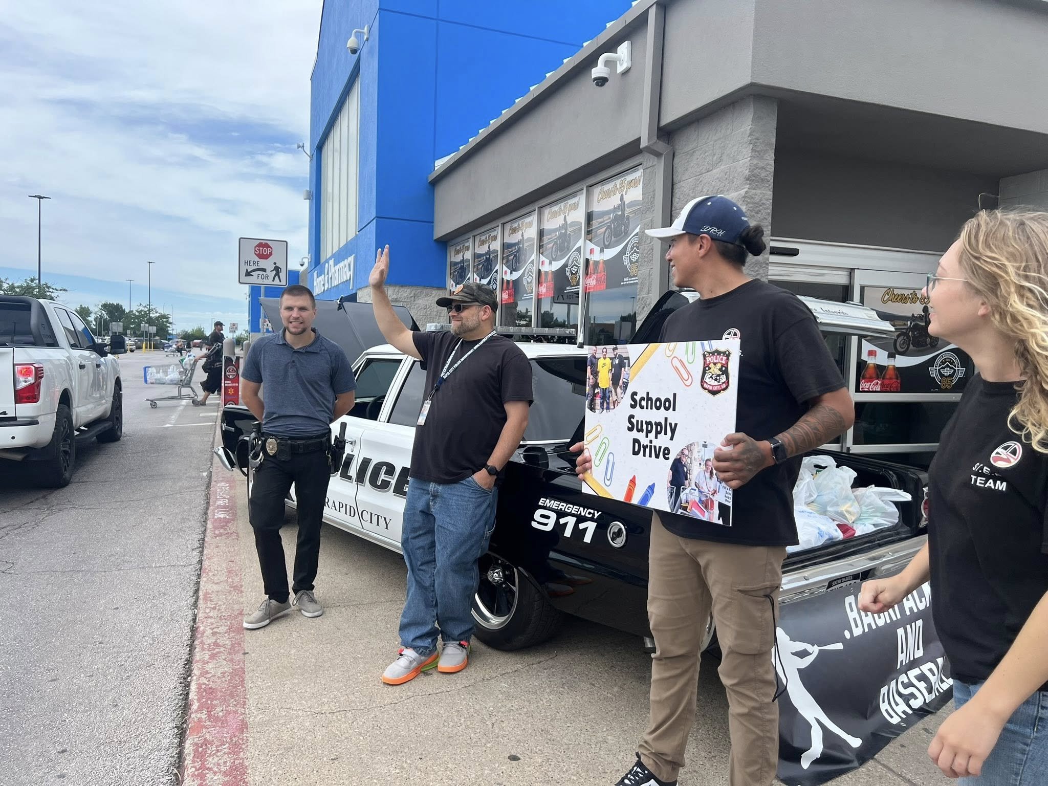 People participating in a school supply drive outside a store, with police officers, a man holding a sign, and a woman near a police car with bags of supplies in the truck bed.