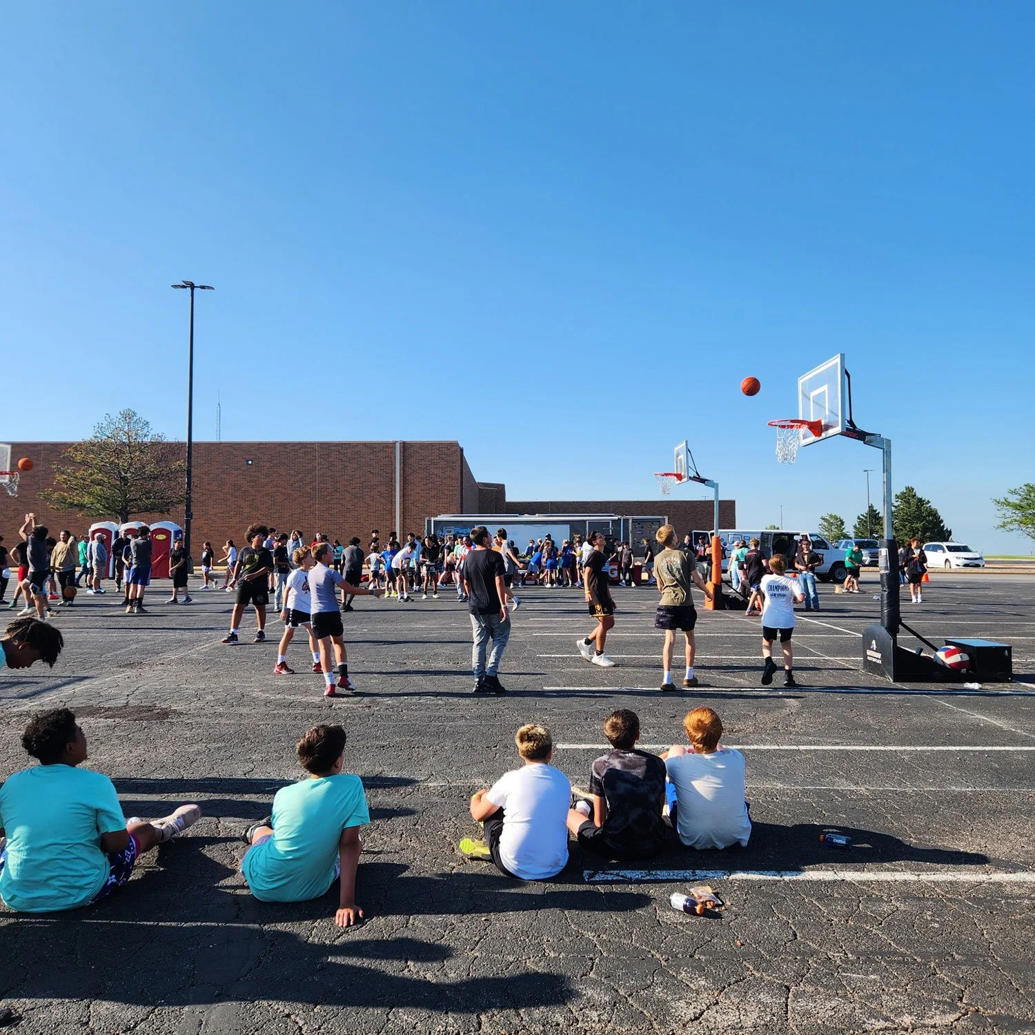 Group of children playing basketball on an outdoor court with a large crowd in the background under a clear blue sky.