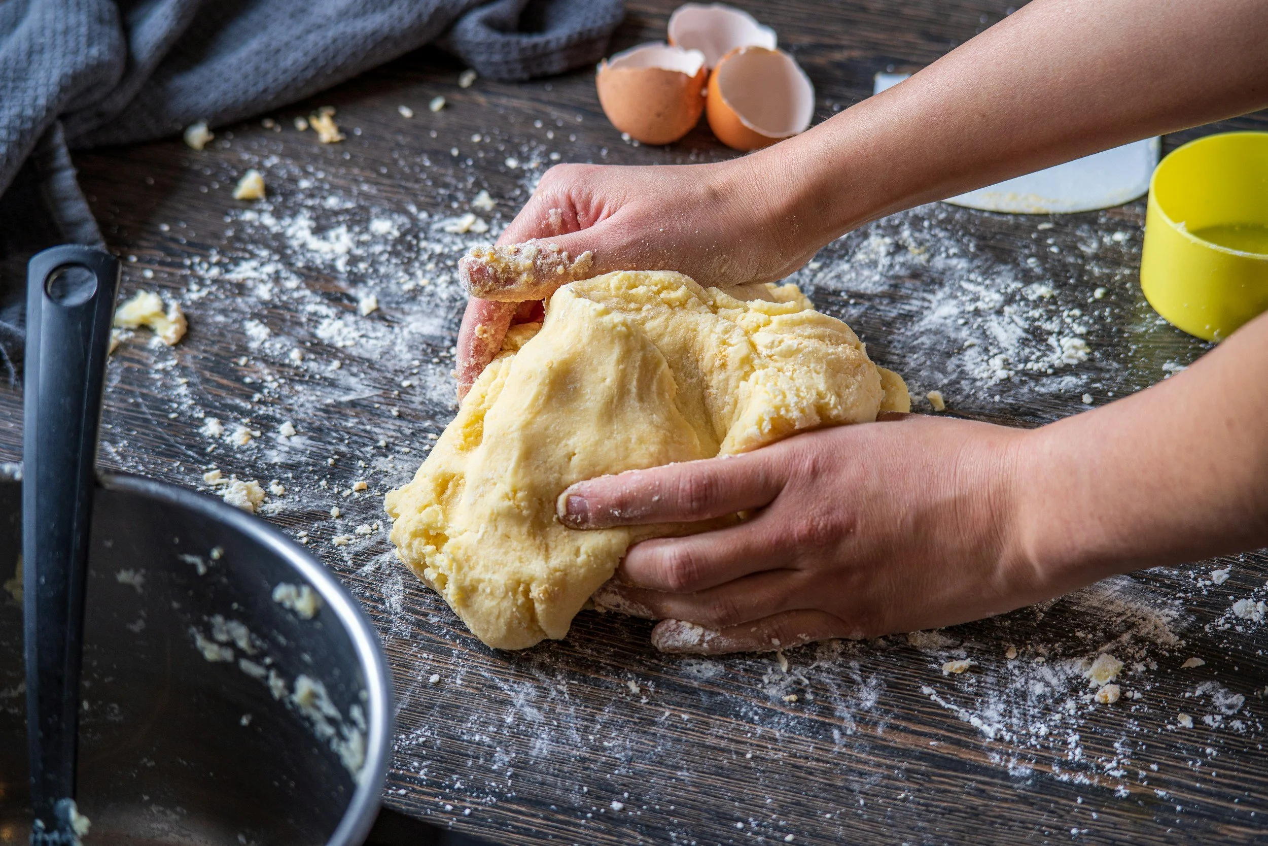 Person kneading dough on a dark wooden surface with eggshells, flour, and baking tools around.