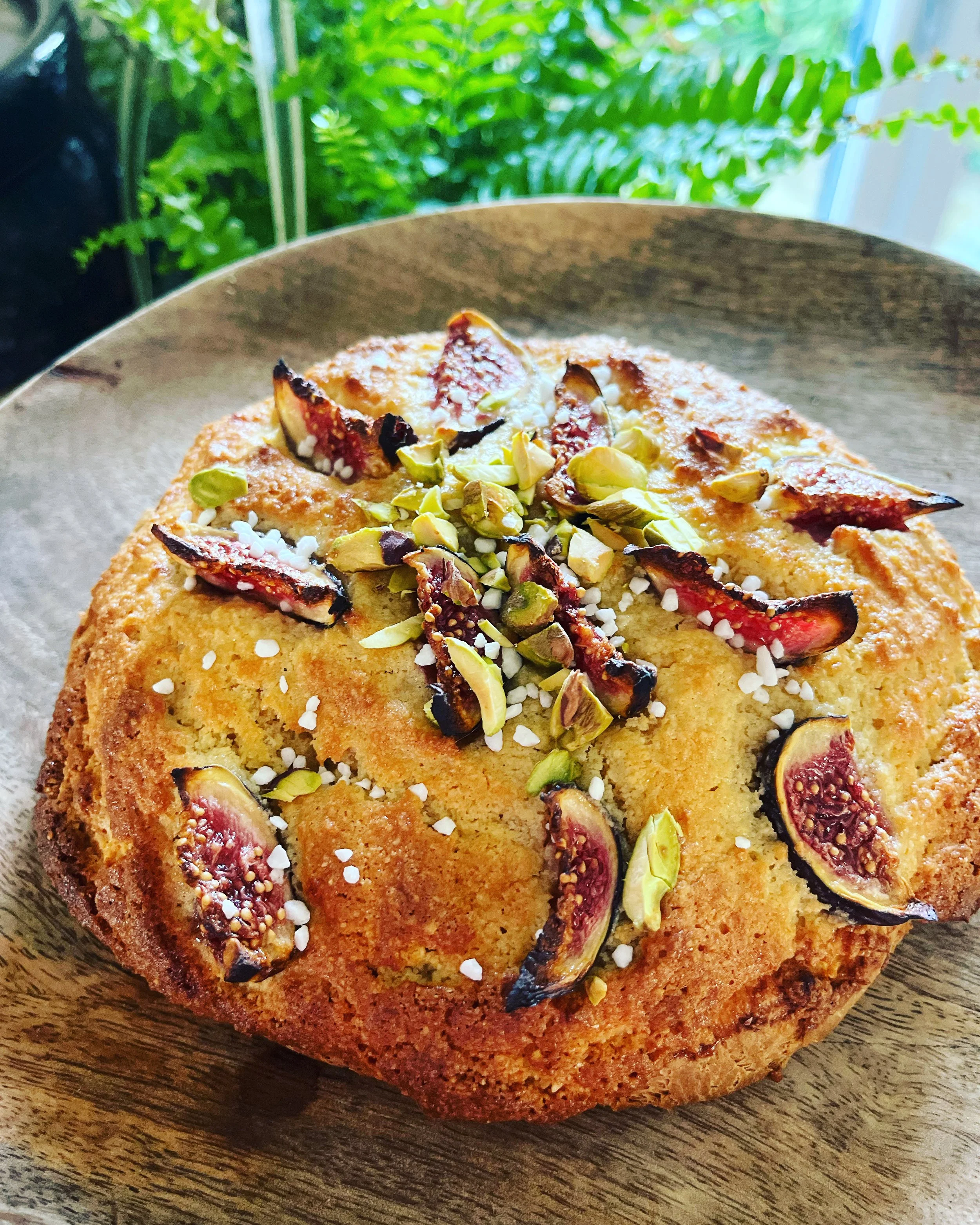 A close-up of a round cake topped with sliced figs, chopped pistachios, and coarse salt on a wooden plate, with green plants and bright sunlight in the background.