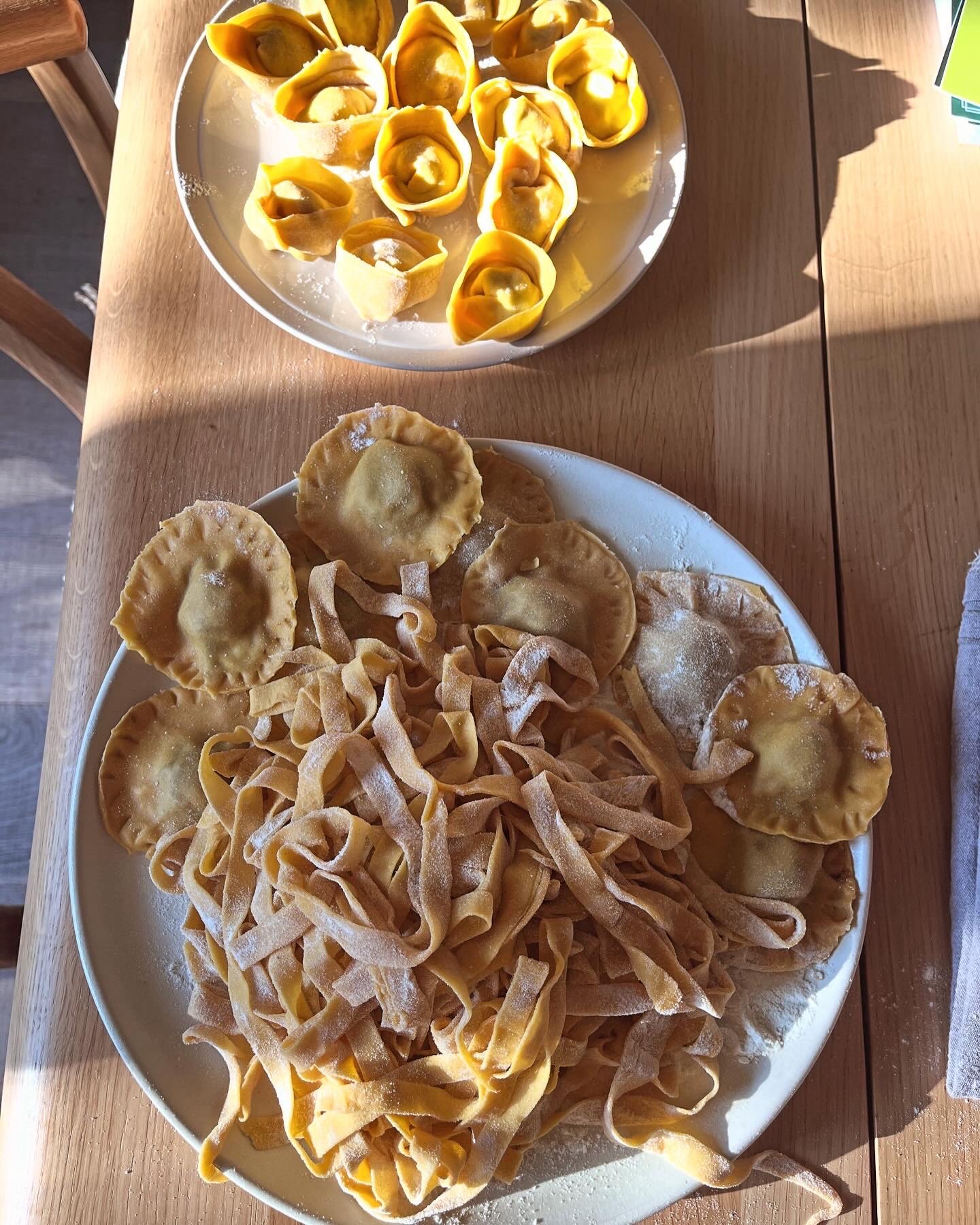 Plate of freshly made ravioli pasta and a smaller plate of uncooked tortellini pasta on a wooden table.