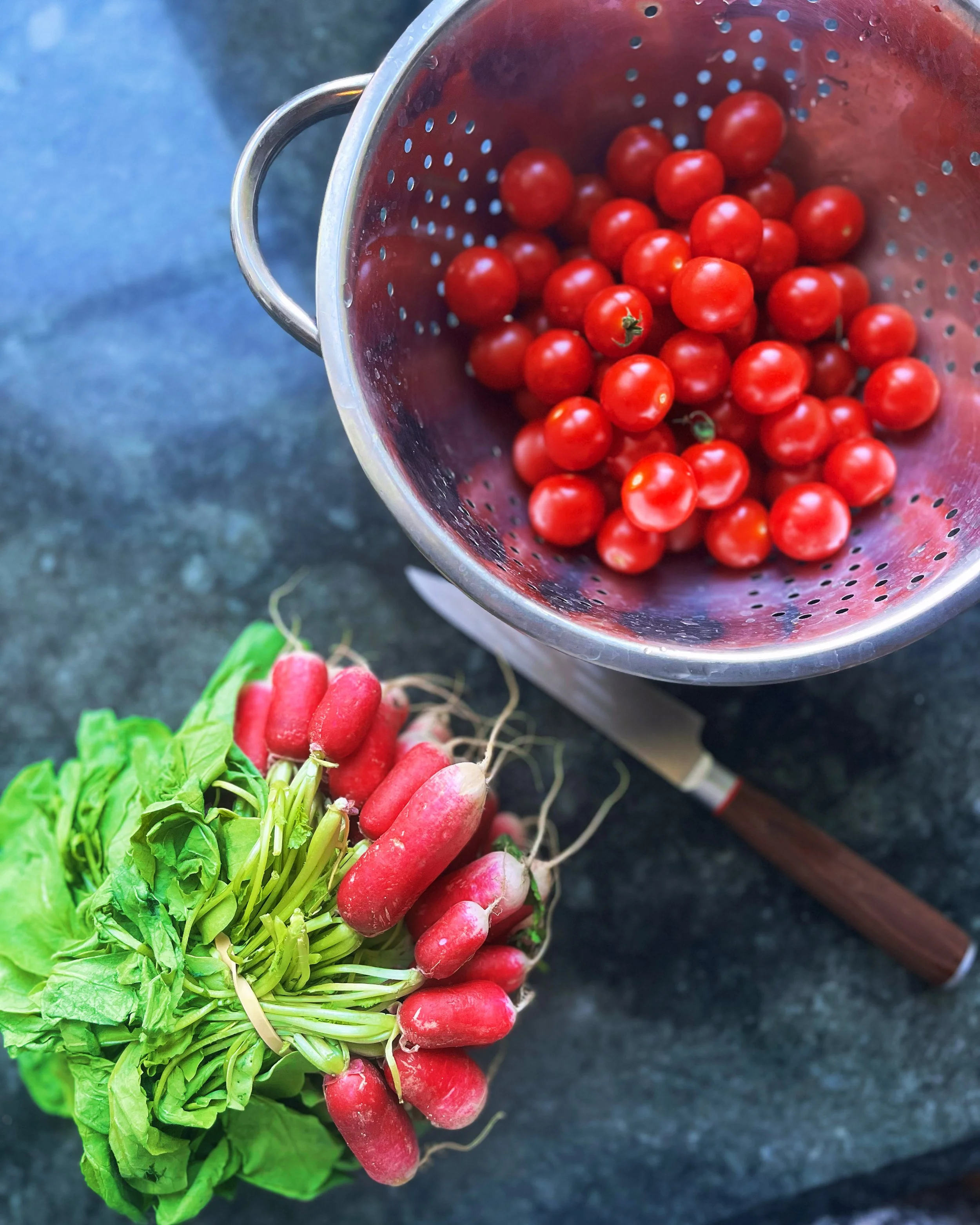 A bunch of fresh radishes with green leaves and roots, placed next to a stainless steel colander filled with red cherry tomatoes on a dark countertop.