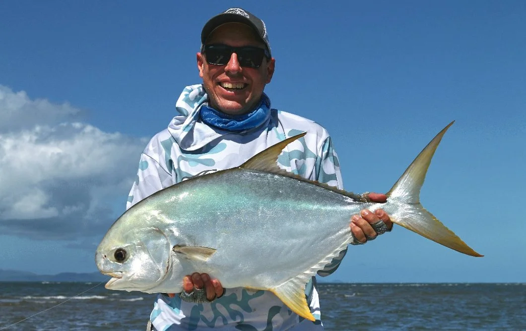 A man wearing sunglasses, a cap, and a white camo shirt holding a large fish with a grayish body and yellow fins at the beach.