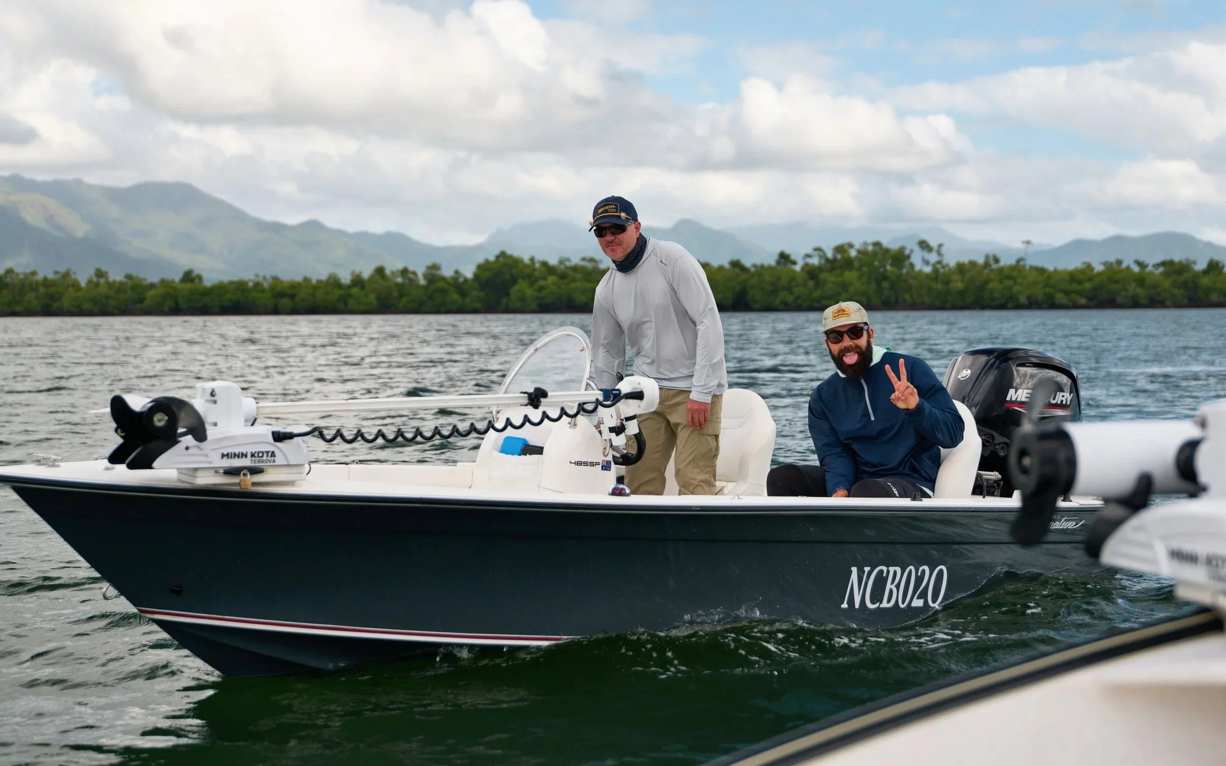 Two men on a small boat with outboard motor on a body of water, with mountains and a partly cloudy sky in the background. One man is standing and the other is sitting, making a peace sign.