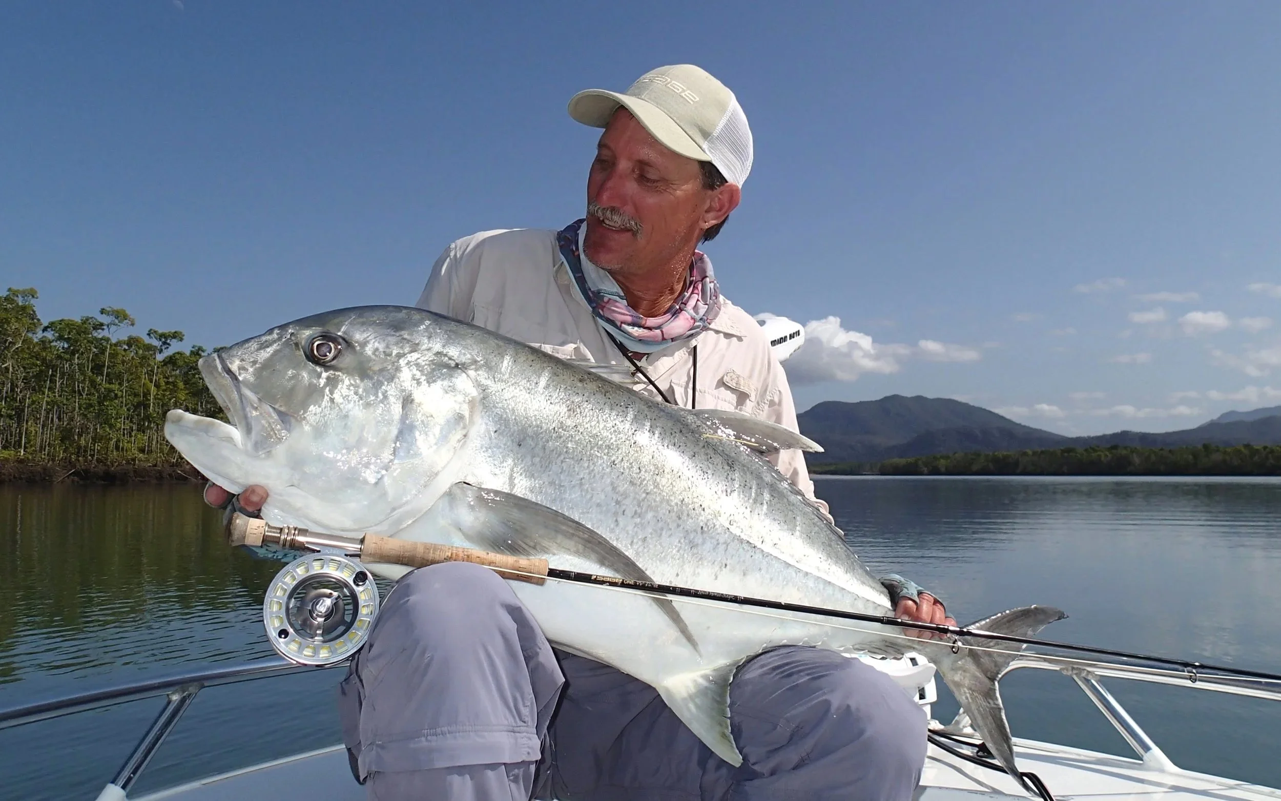 An older man in outdoor fishing gear holding a large fish on a boat with water and mountains in the background.