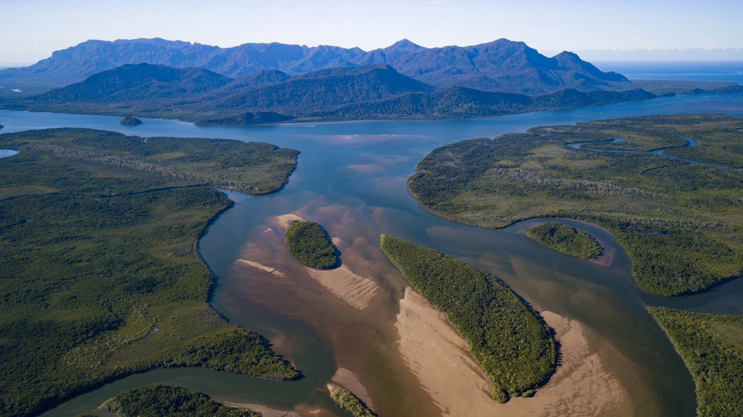 Aerial view of a winding river with land islands and green forests, mountains in the background, and blue sky.