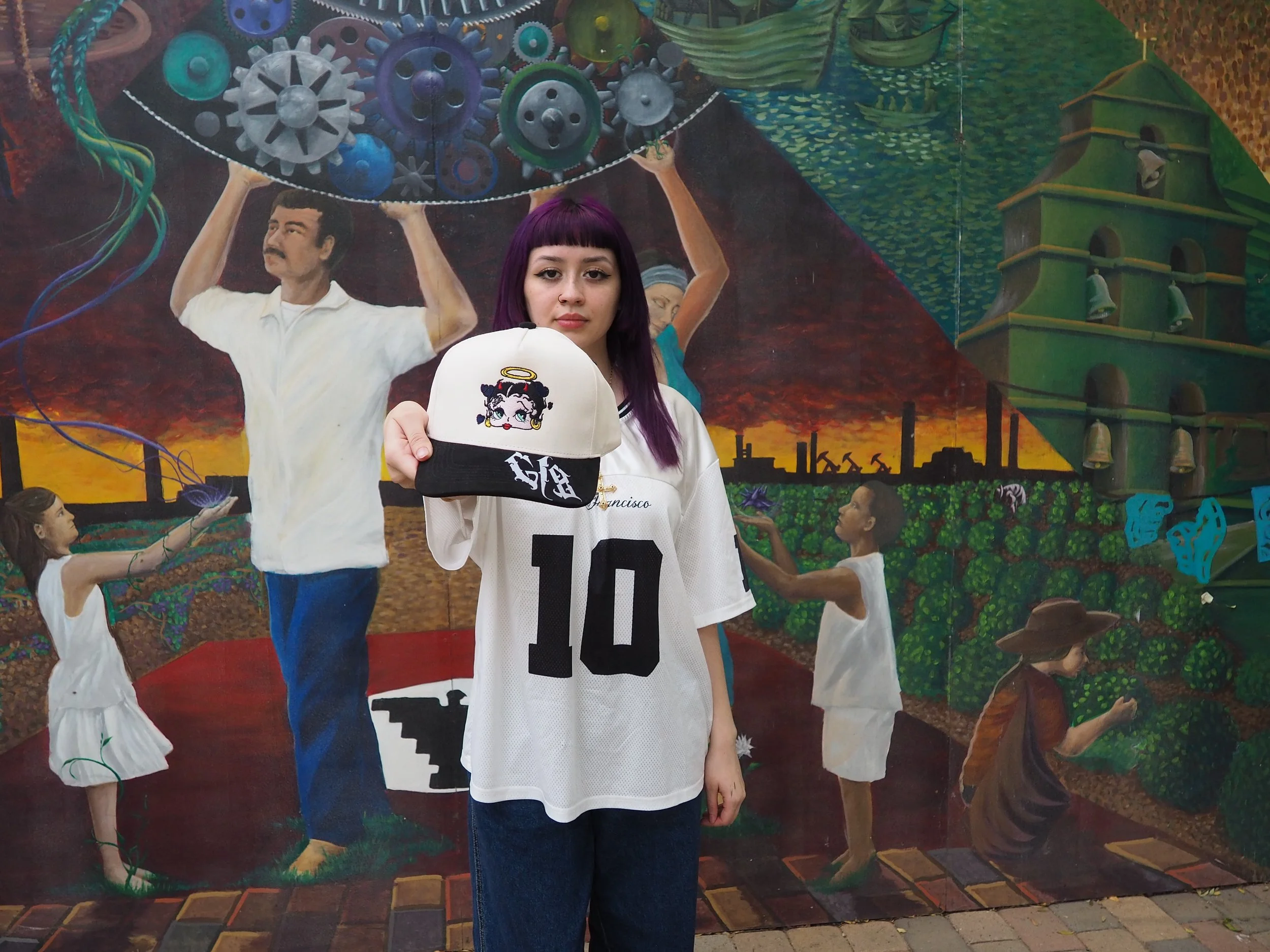 A young woman with purple hair wearing a white sports jersey with the number 10 and holding a beige and black cap with Betty Boop artwork, standing in front of a colorful mural featuring people, gears, and buildings.