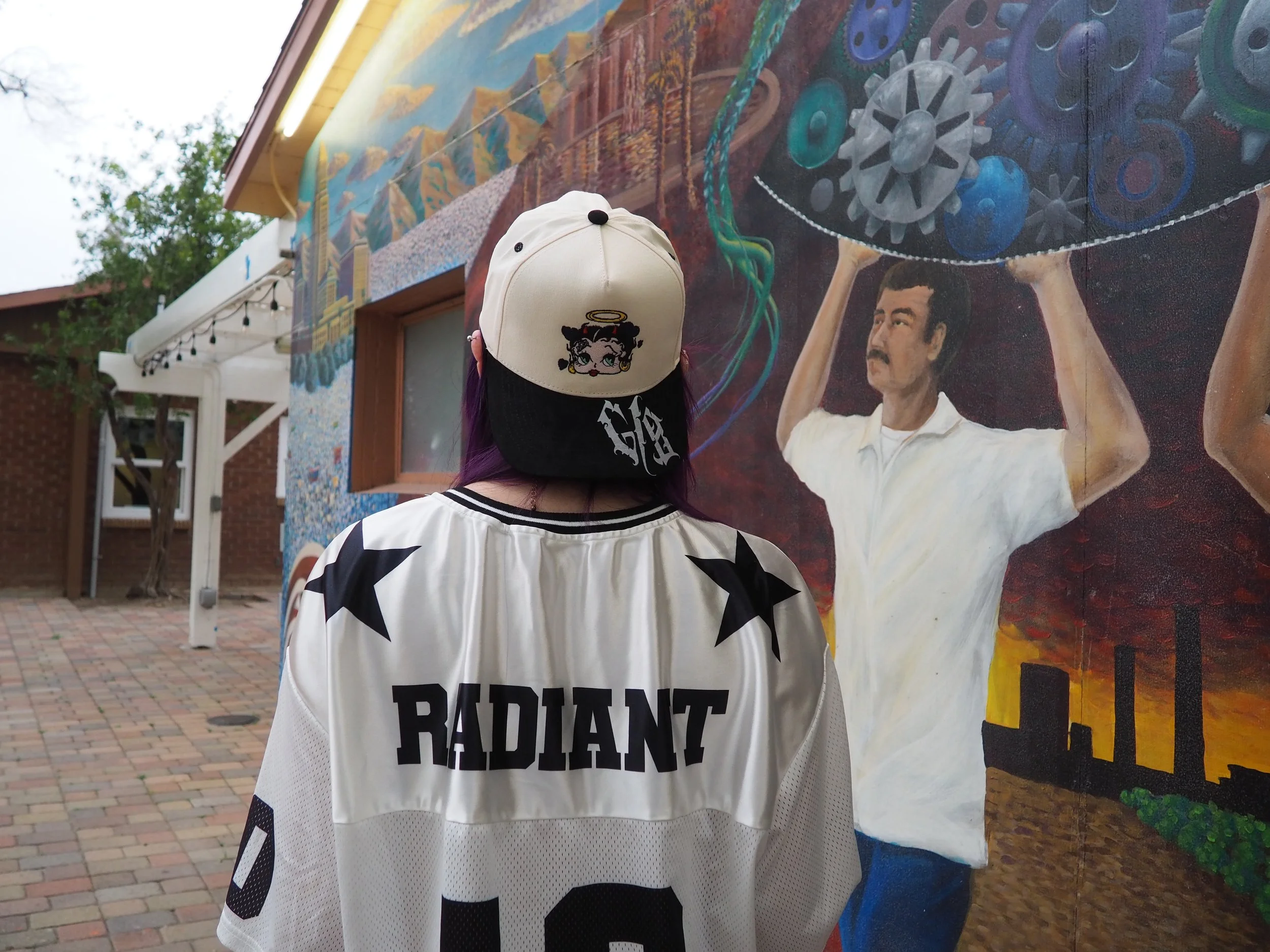 Person with purple hair wearing a white baseball cap with Betty Boop on it and a white sports jersey with black stars and the word 'RADIANT' on the back, standing in front of a mural depicting a man holding a large gear in a cityscape at sunset.