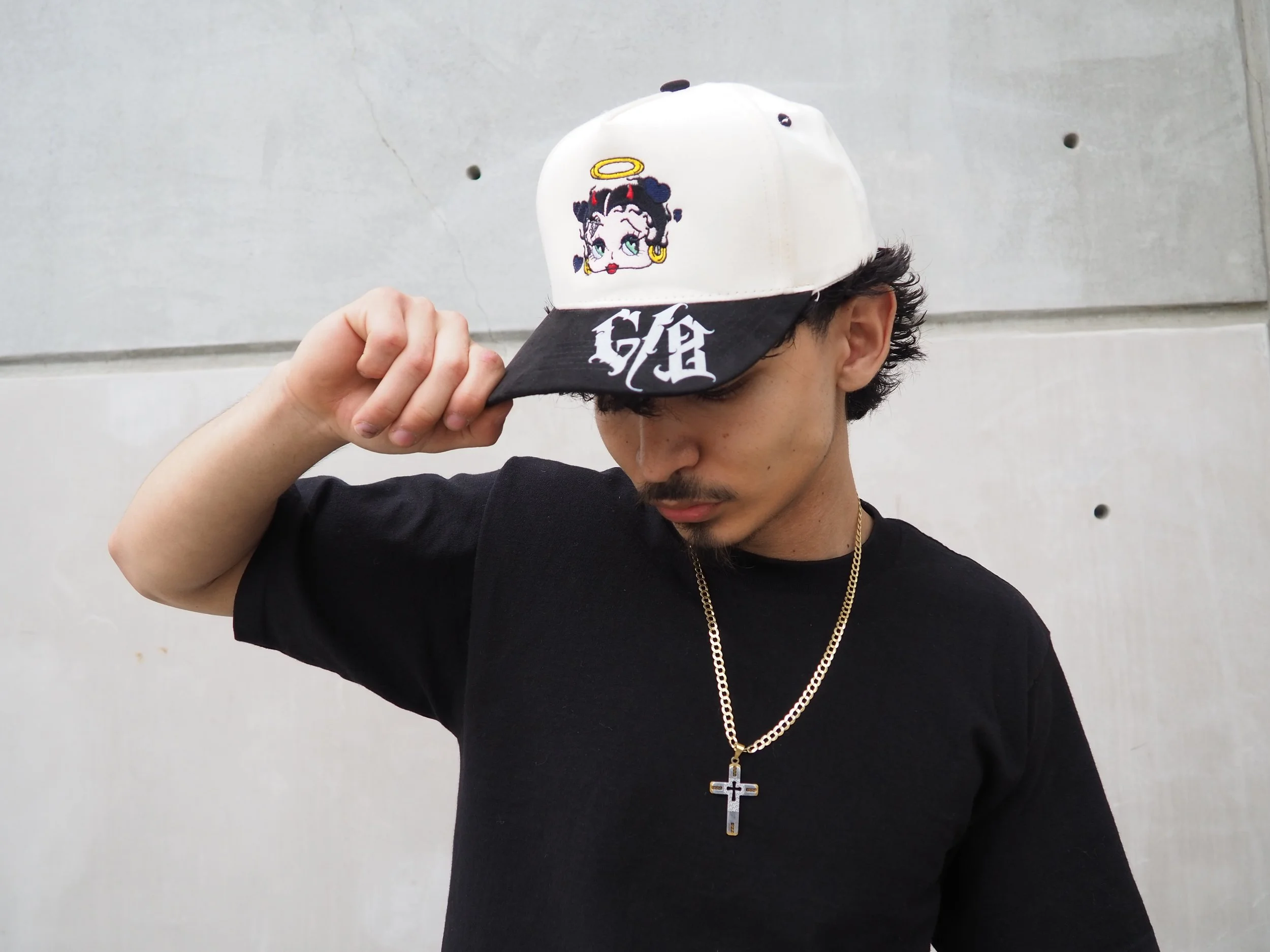 Young man wearing a black t-shirt, gold chain with a cross pendant, and a white cap with Betty Boop graphic and black brim, standing against a concrete wall.