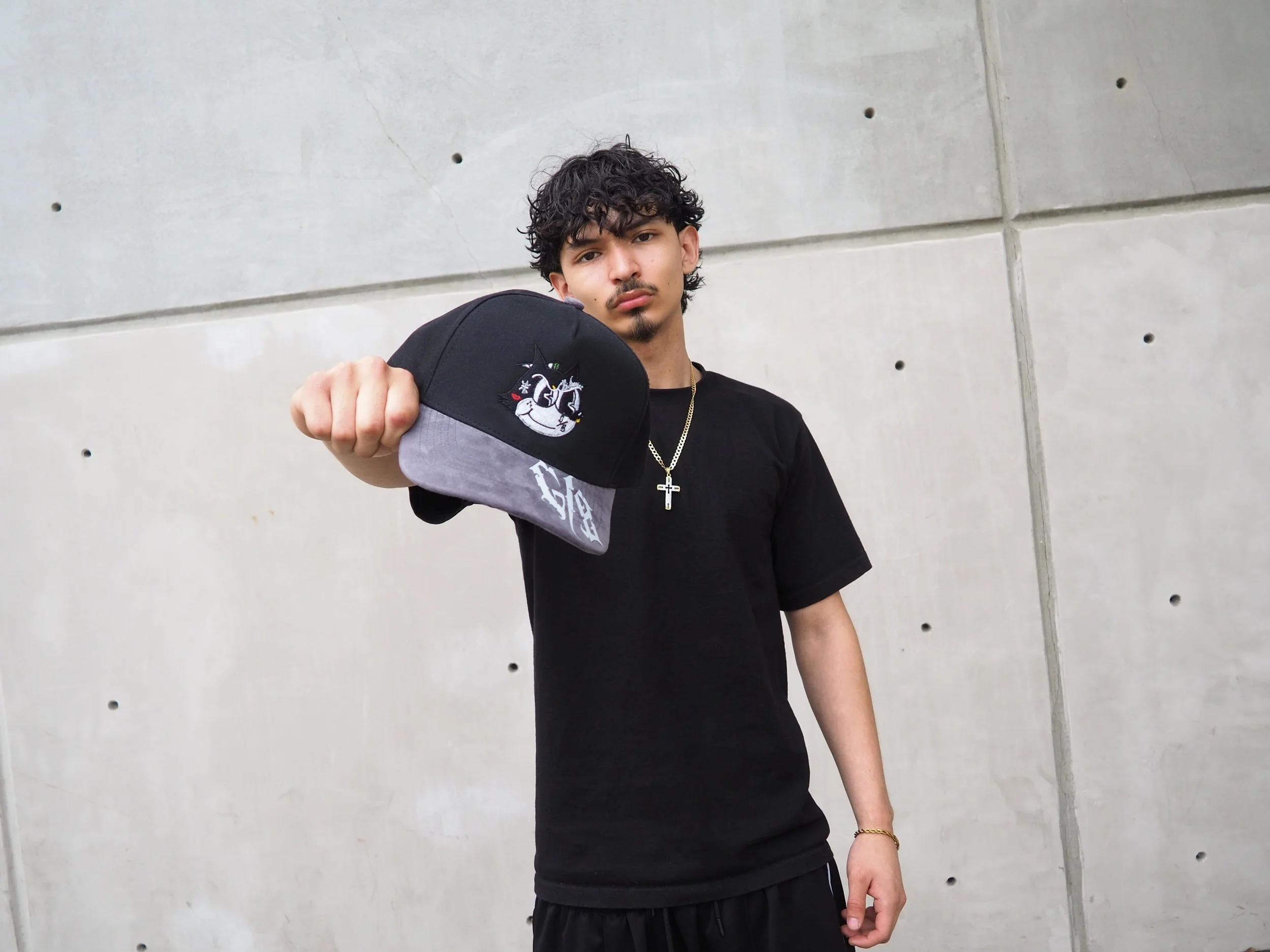 Young man with curly black hair and a black t-shirt holding a black and gray cap with cartoon character embroidered on it, standing against a concrete wall.