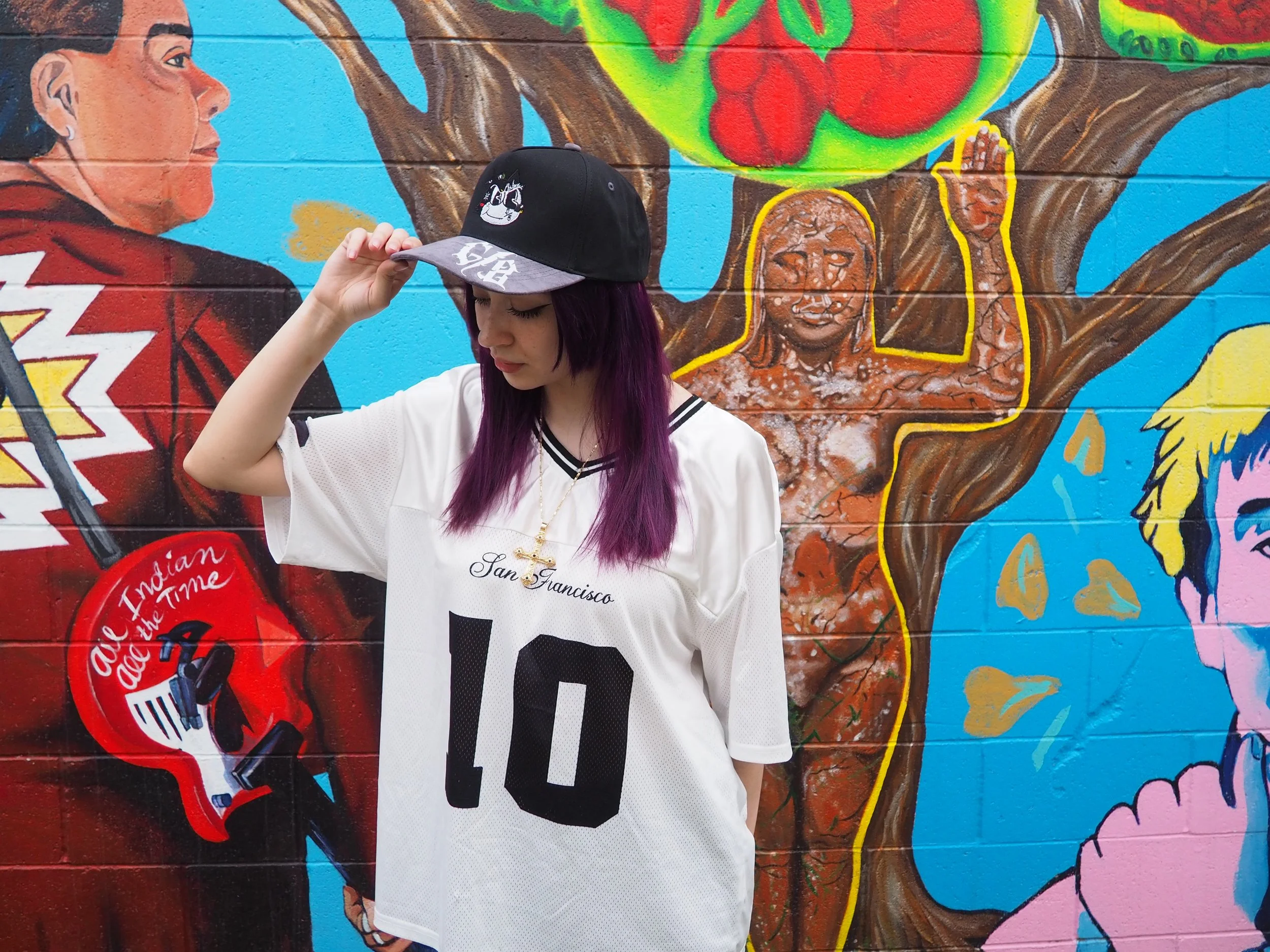 A young woman with purple hair and a black cap wearing a white jersey with the number 10 stands in front of a colorful mural painted on a brick wall. The mural features various figures, including a person holding a guitar with the words "All Indian A