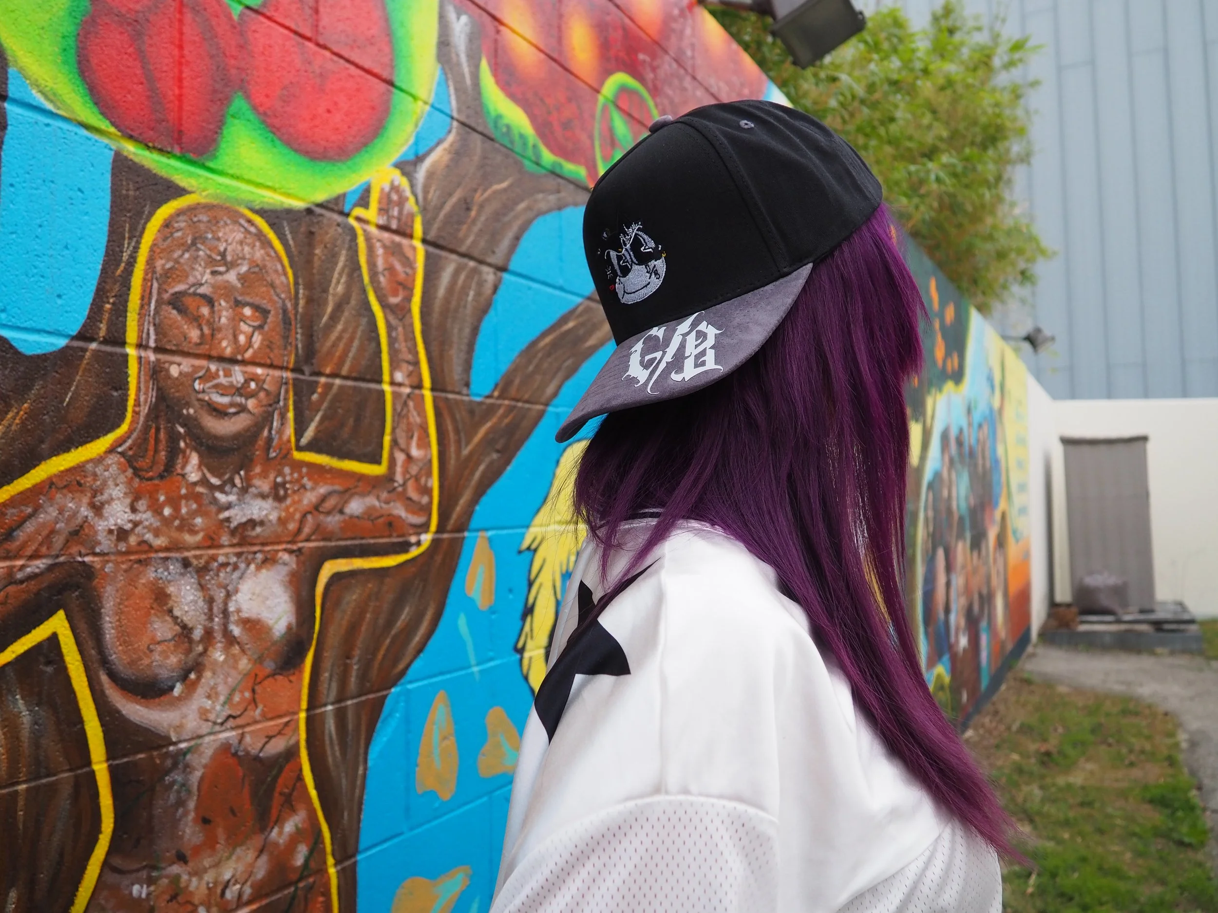 Person with purple hair wearing a black and gray baseball cap and white shirt, looking at a colorful mural on a brick wall featuring a woman and trees.