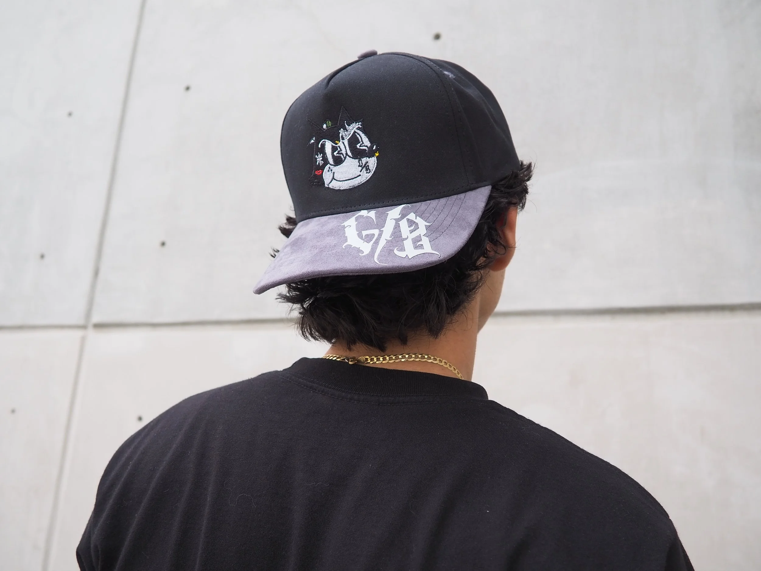 Back of a person wearing a black baseball cap with a cartoon unicorn on the front and the word 'GIA' written on the brim, and a black t-shirt with a gold chain necklace.