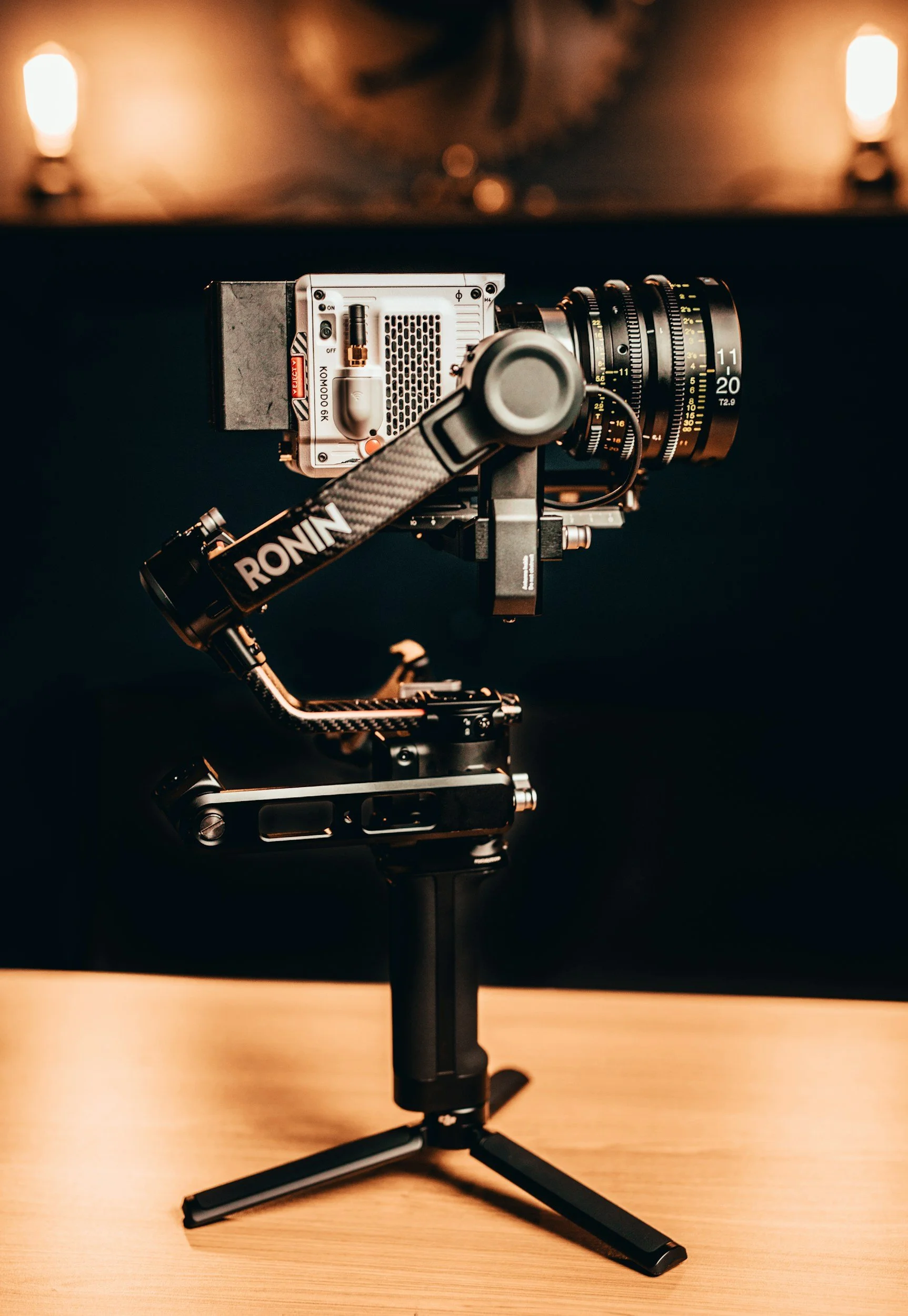 A professional camera mounted on a gimbal stabilizer on a wooden surface, with blurred warm lights in the background.