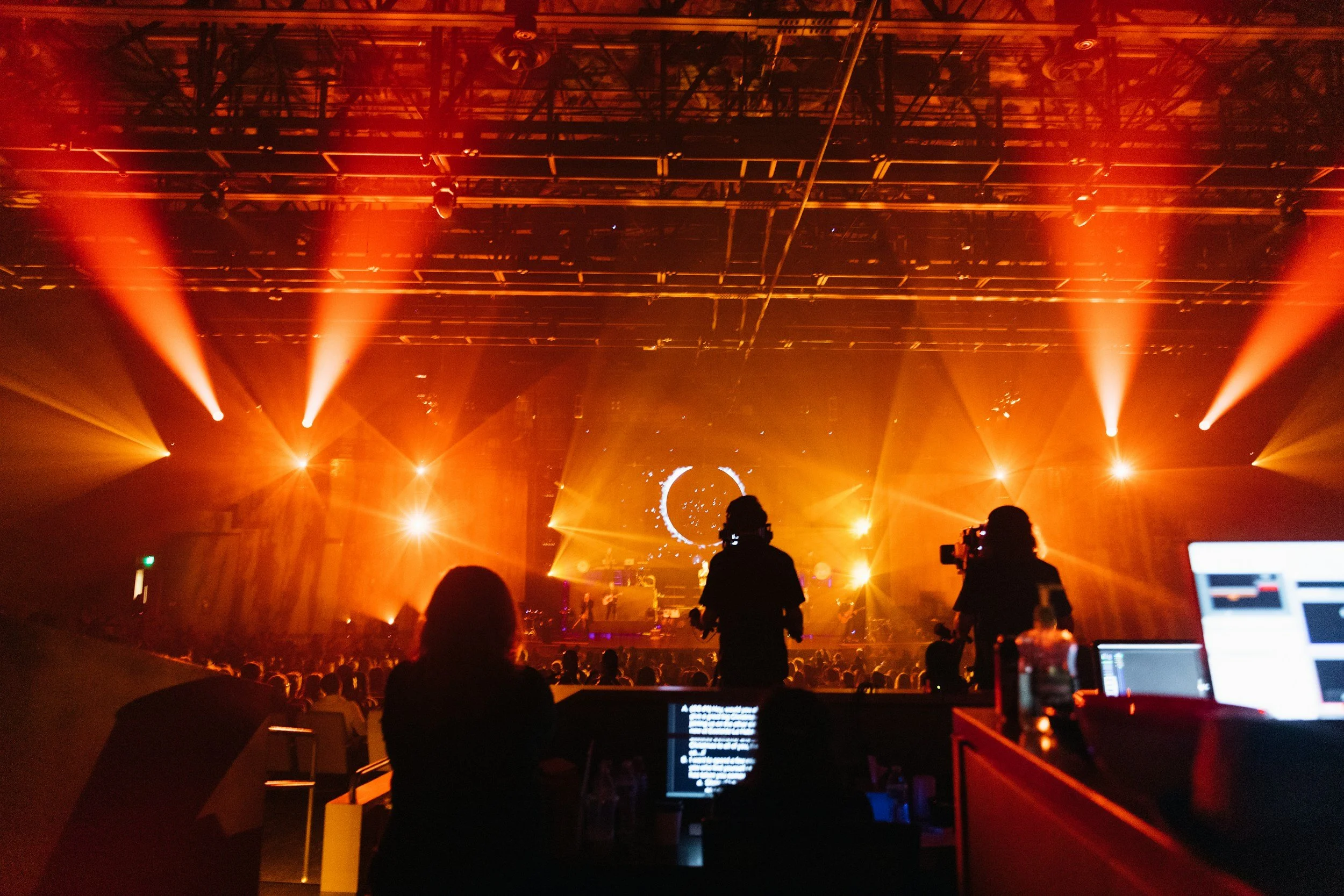 Silhouettes of people, likely at a concert or festival, in front of a stage with bright orange and yellow lights, a large circular screen with visuals, and a crowd of audience members.