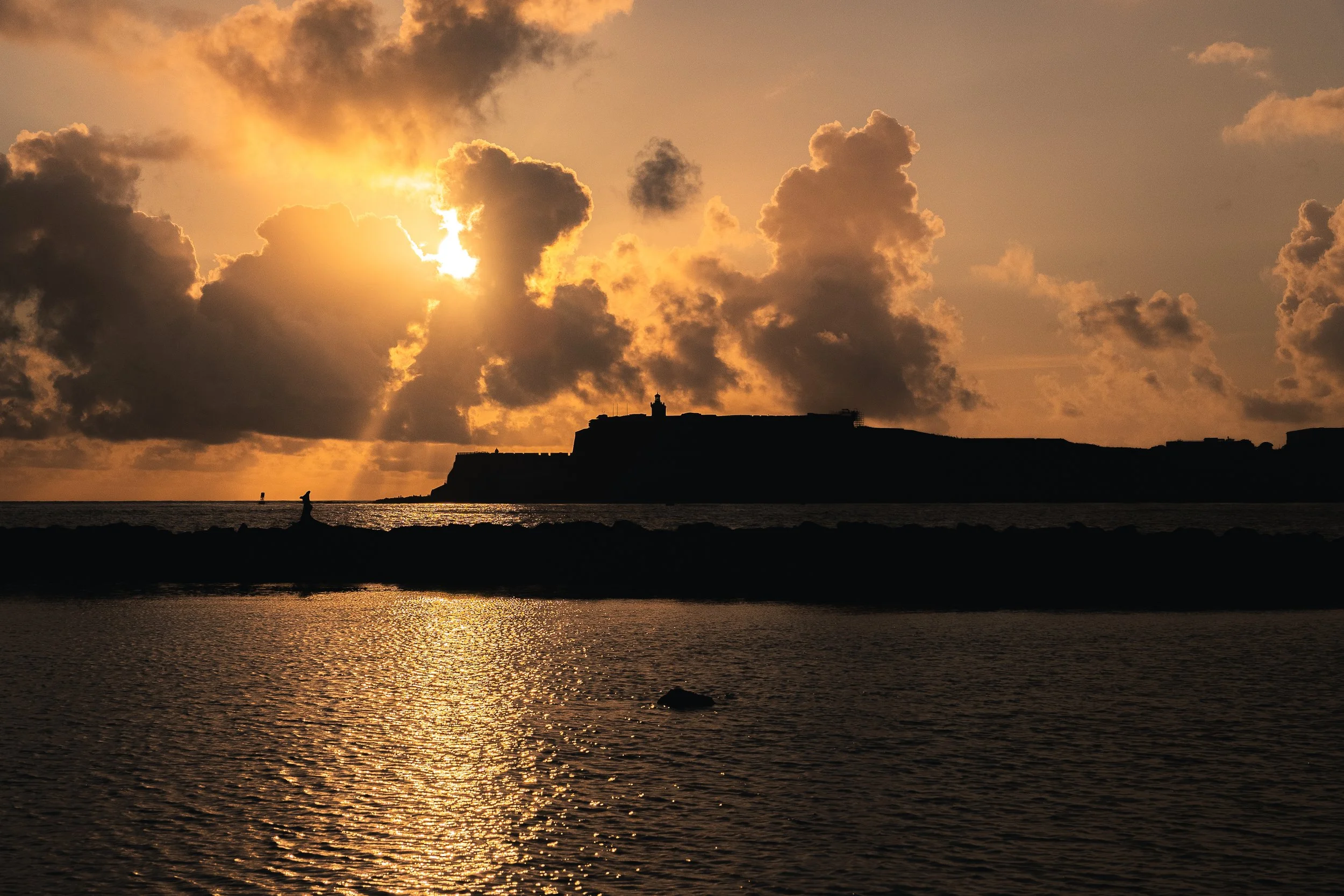 A silhouette of a distant fort or castle on a hill is visible against a sunset sky with dramatic clouds, reflecting on the water below, in a coastal scene.