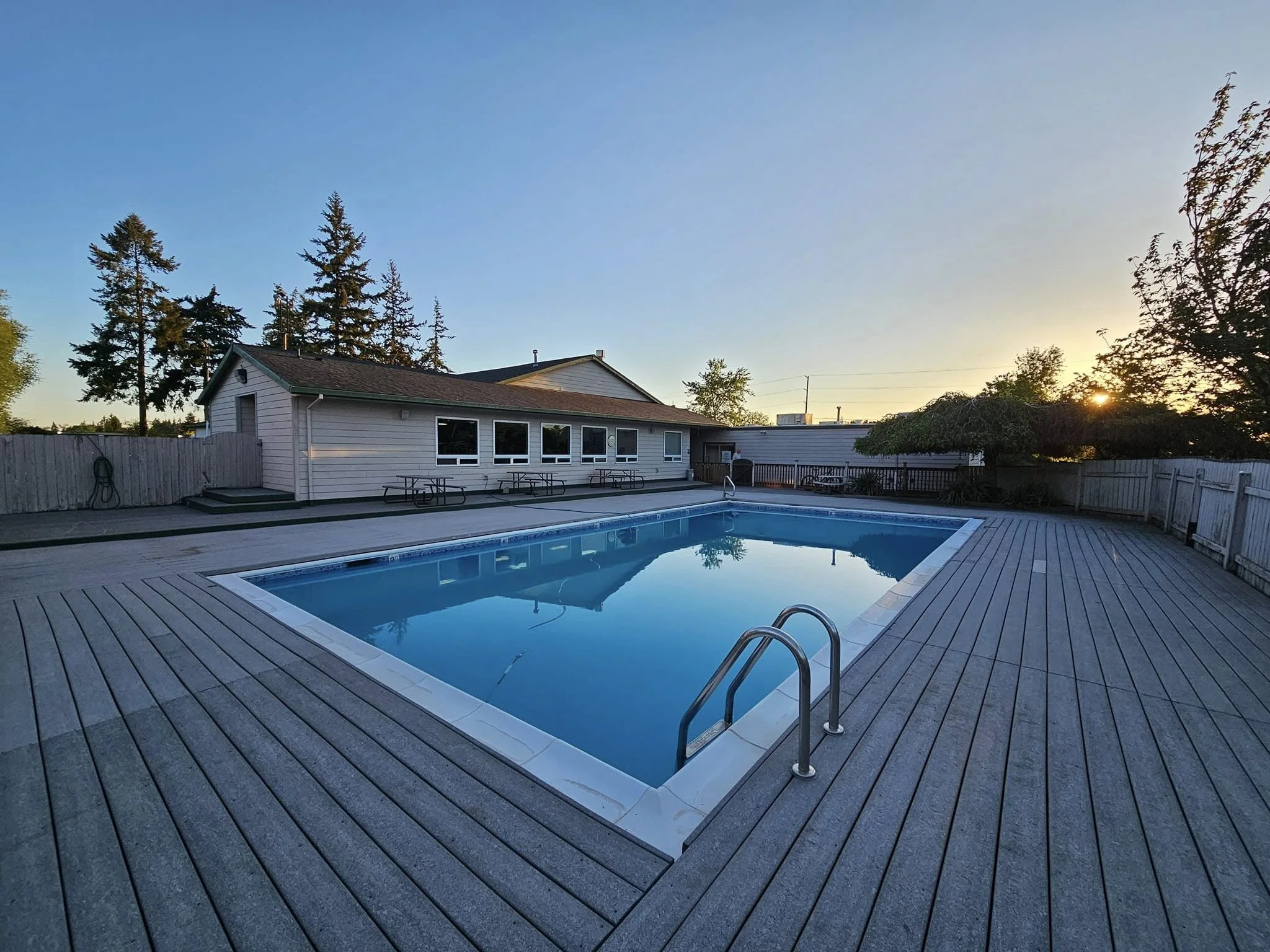An outdoor swimming pool with a wooden deck, surrounded by a fence, in front of a house with several windows, during sunset.