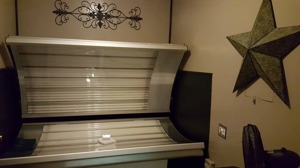 Room with beige walls decorated with a large metal wall art piece, a big 3D star decoration, a small framed picture, and a black fan. There are two windows with closed blinds and a small picture frame on a dark surface.