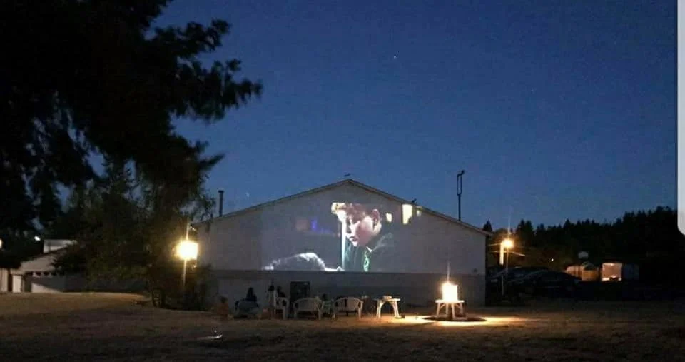 An outdoor movie screen on the side of a building shows a scene from a film with a young boy. Chairs and tables are set up in front of the screen on a grassy area, with outdoor lighting illuminating the scene during what appears to be nighttime.