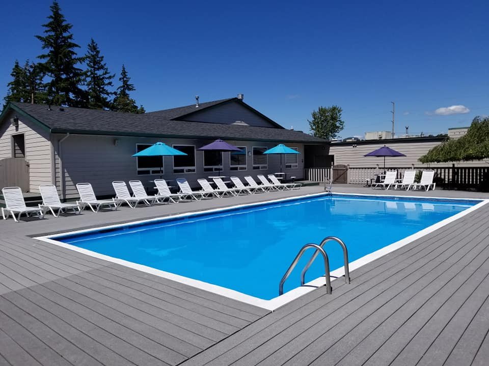 An empty swimming pool with a gray deck, surrounded by white lounge chairs and colored umbrellas, outside a single-story building under a blue sky.