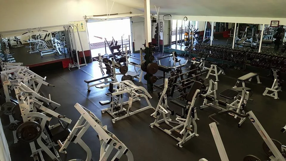 Empty gym with weightlifting equipment including benches, dumbbells, barbells, and machines in a spacious room with large mirrors.