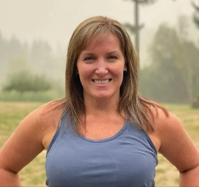 A woman outdoors smiling, wearing a blue tank top, with trees and a cloudy sky in the background.