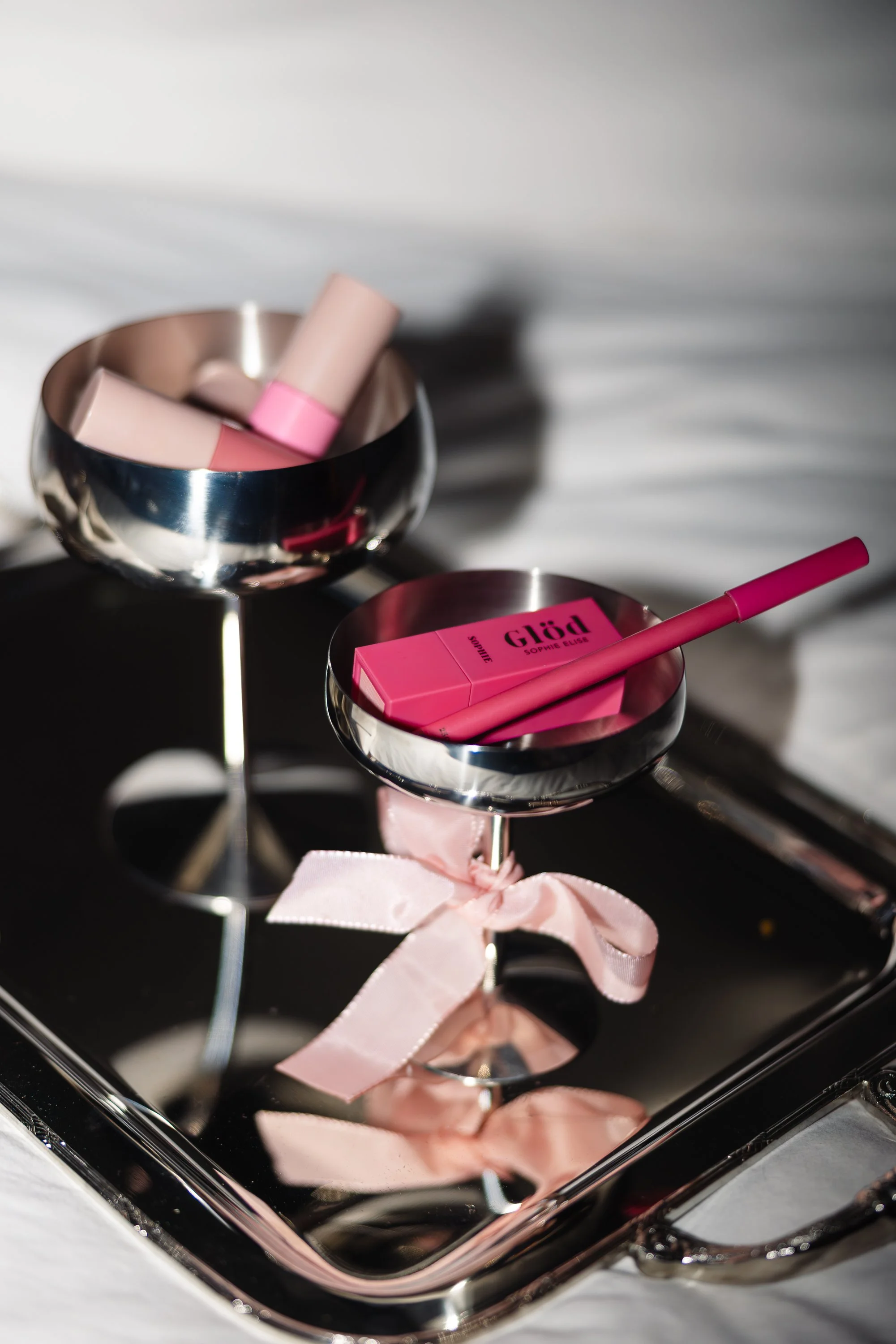 Beauty products including pink and beige lipsticks and a pink package labeled 'Glam' on a reflective silver tray with a pink ribbon.