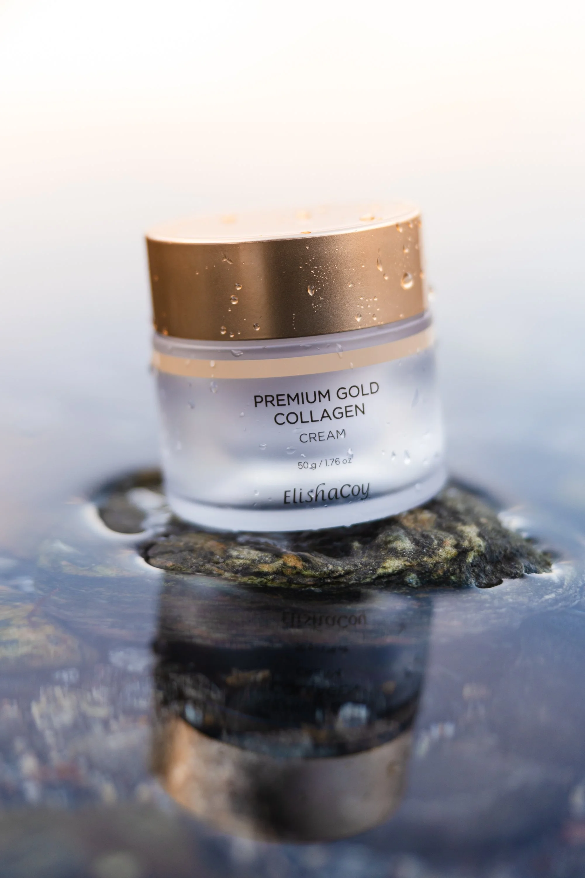 A jar of premium gold collagen cream sitting on a rock in water with a reflection underneath.