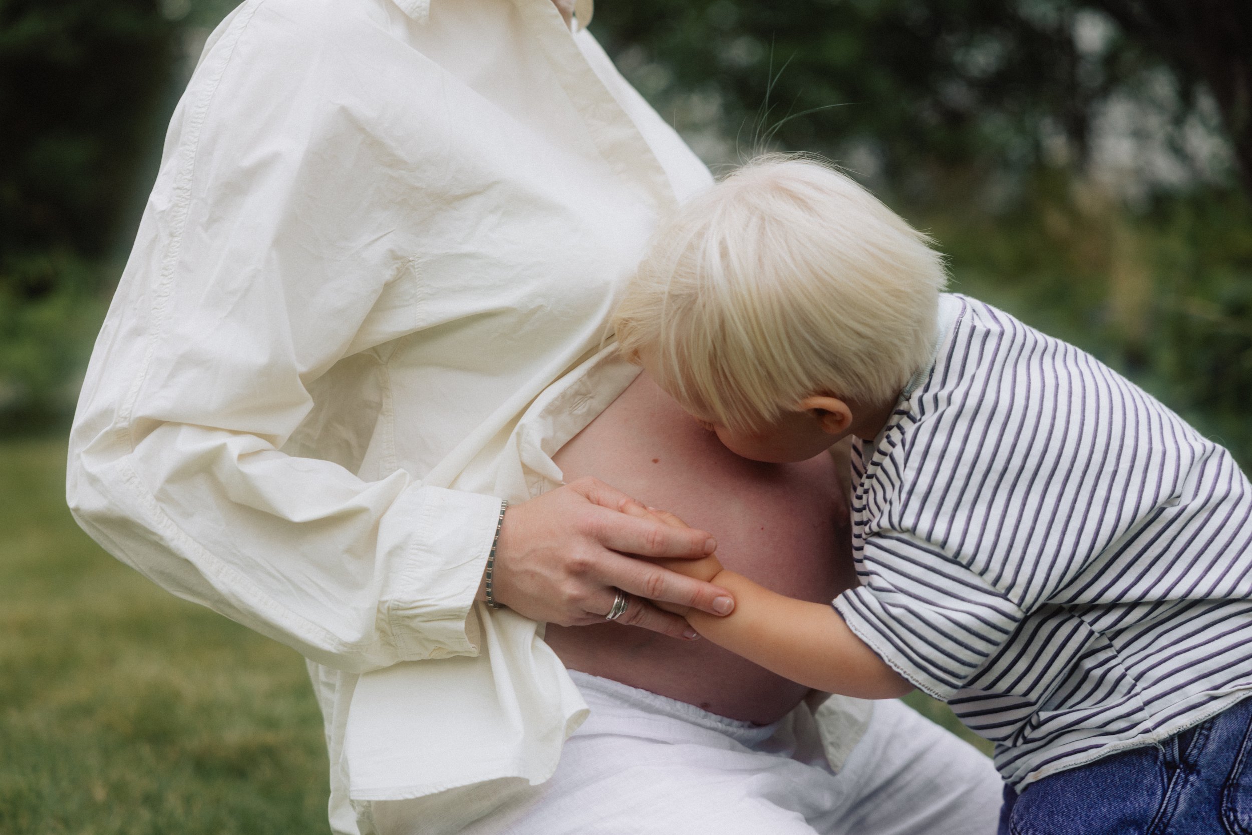 En person som bærer en gravid mage mens et barn kysser den på magen utendørs.