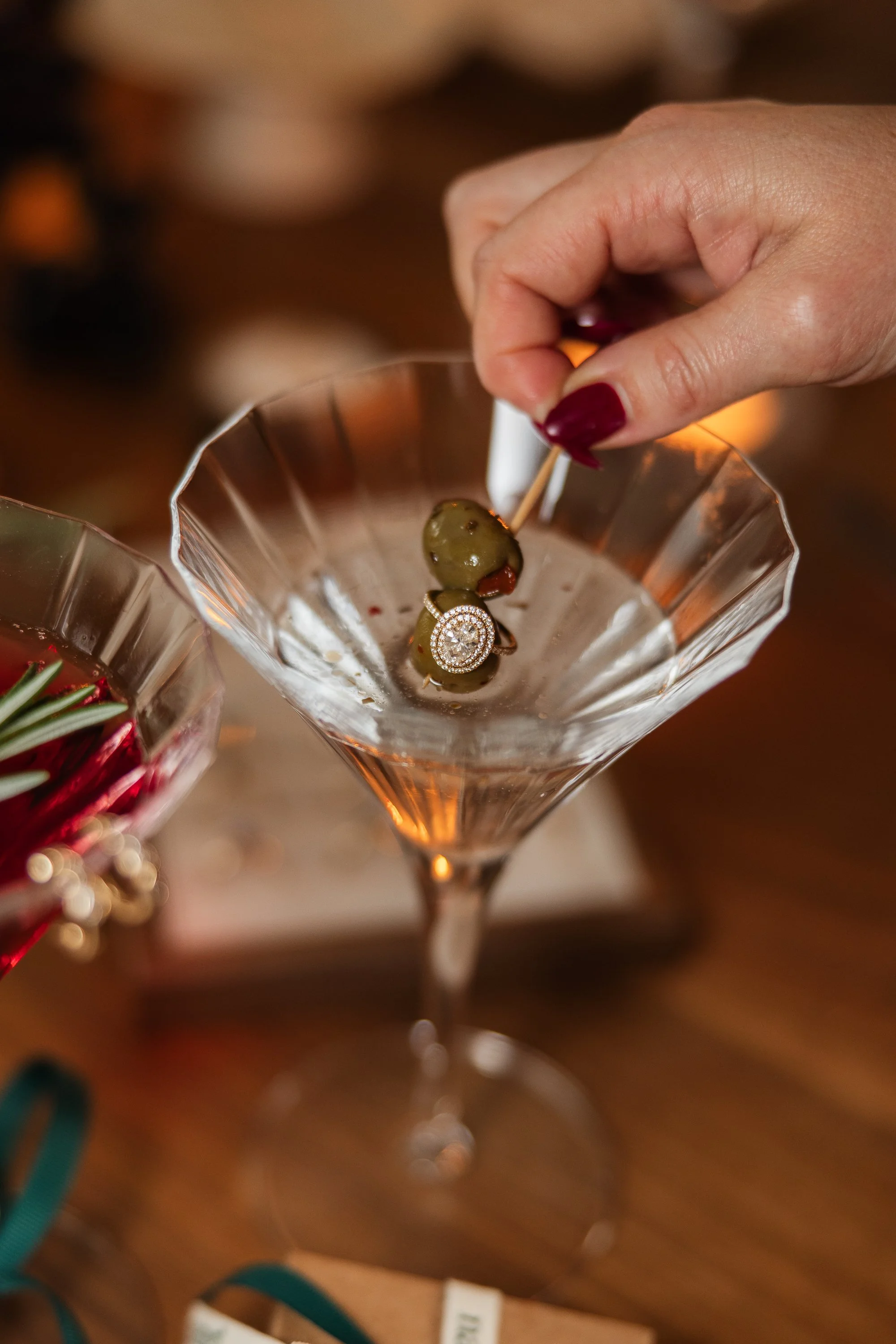 A hand with red painted nails holding a toothpick with a green olive garnished with a small decorative piece, resting over a cocktail glass